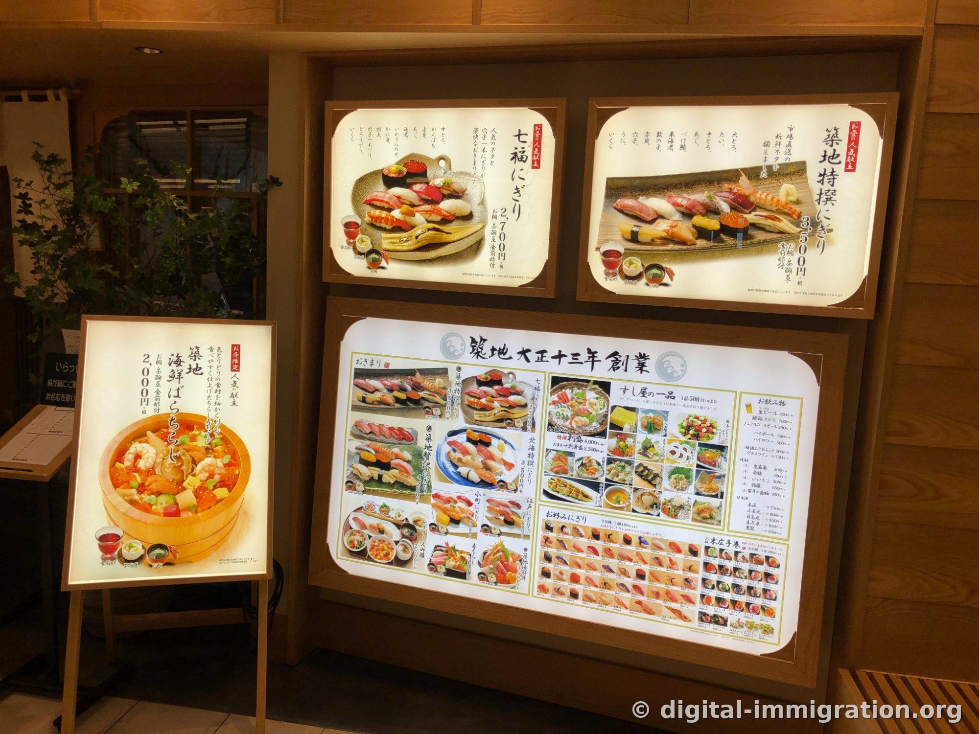Panneaux de menu lumineux à l'entrée du restaurant. On voit le七福にぎり à 2 700 yens, le築地特撰にぎり à 3 500 yens, et sur le chevalet à gauche, le築地海鮮ばらちらし à 2 000 yens