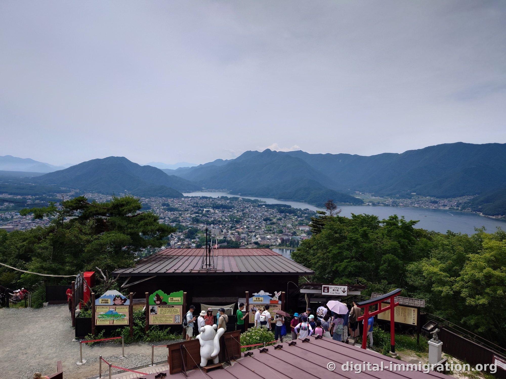 Beyond the summit station roof, Lake Kawaguchiko, the town, and overlapping mountain ridges spread out in a panoramic view, with a red torii gate on the right