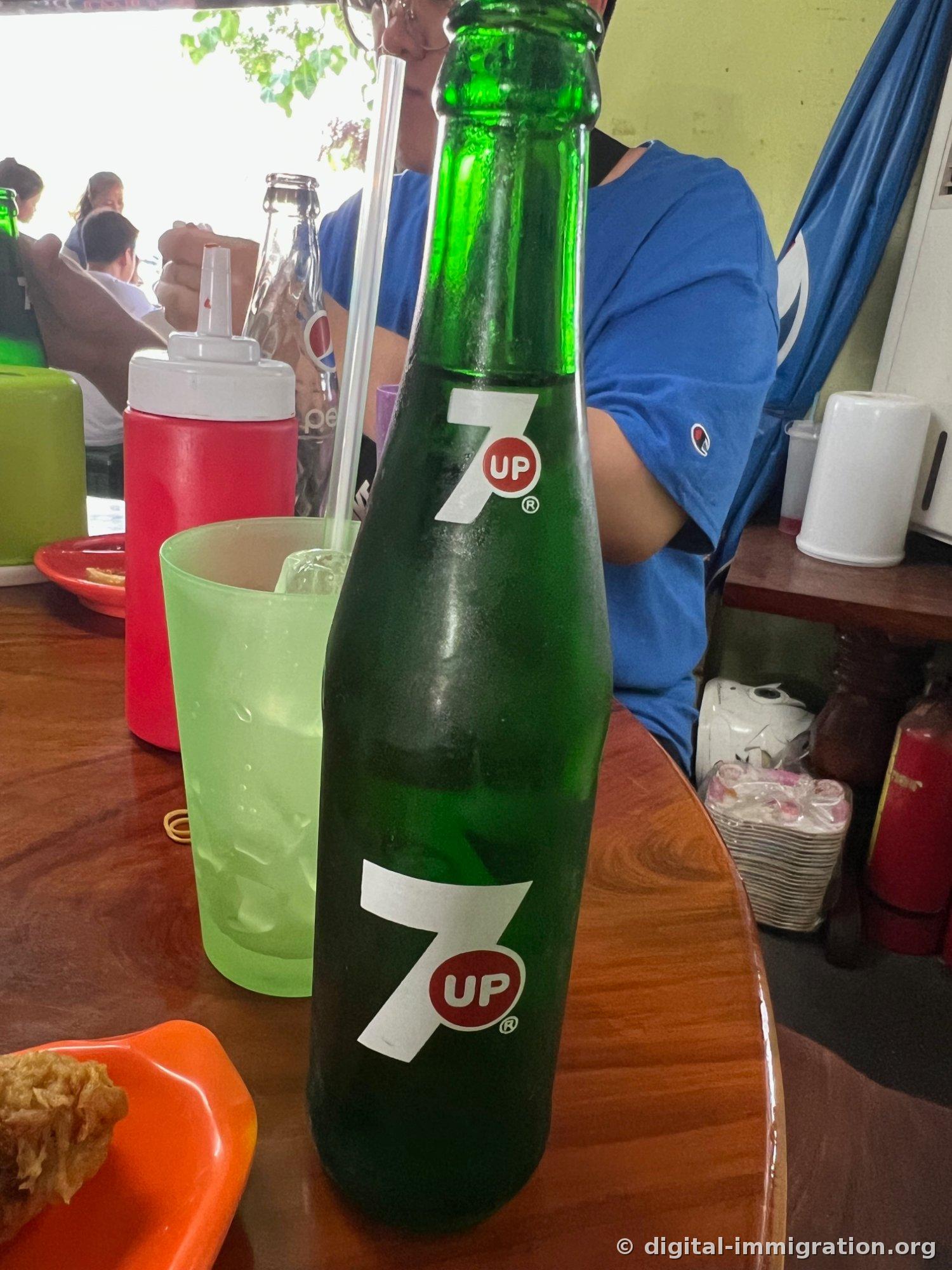 A sweating glass bottle of green Seven Up with a glass straw, next to a pale-green plastic cup of ice and a red sauce container on a restaurant table