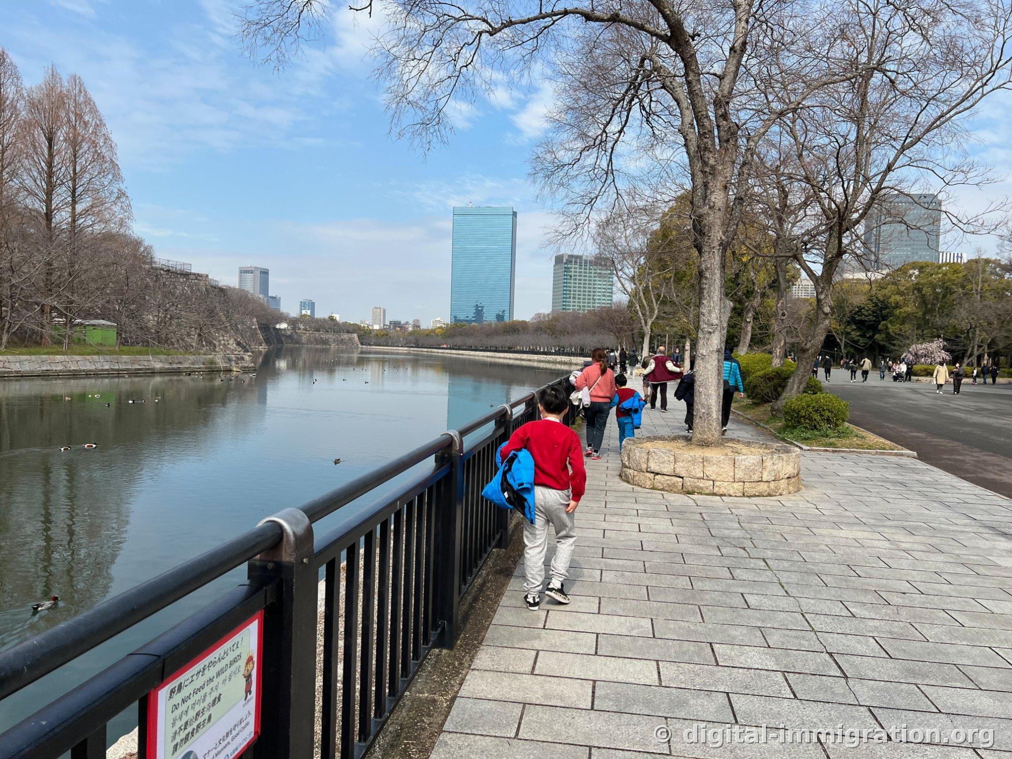 Large allée pavée de pierre longeant le fossé du château d'Osaka. Un enfant ayant enlevé sa veste et sa famille marchent sur le chemin, les gratte-ciel du Osaka Business Park sont visibles en face