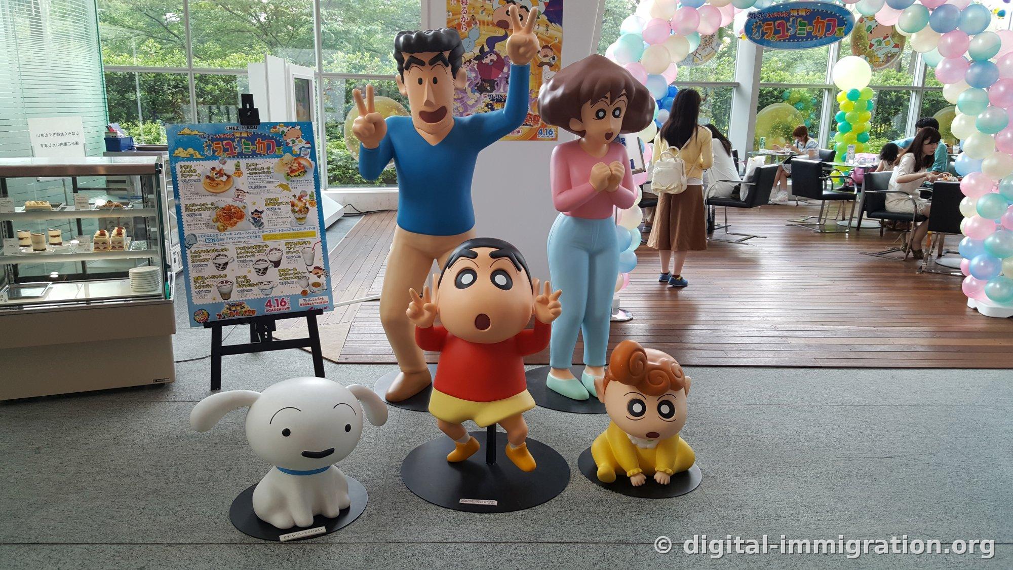 Cinq figurines de la famille de Shin-chan se dressant devant l'entrée du café avec un chevalet-menu de collaboration de café à gauche, une arche de ballons pastel à droite et des clients assis aux tables