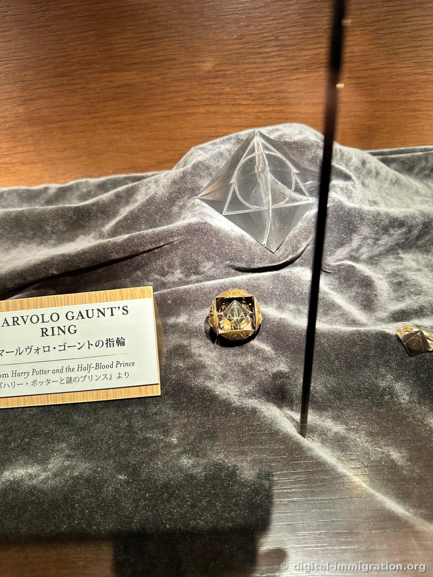 A gold ring and a transparent crystal sculpture engraved with the Deathly Hallows symbol on gray velvet, with the nameplate MARVOLO GAUNT'S RING