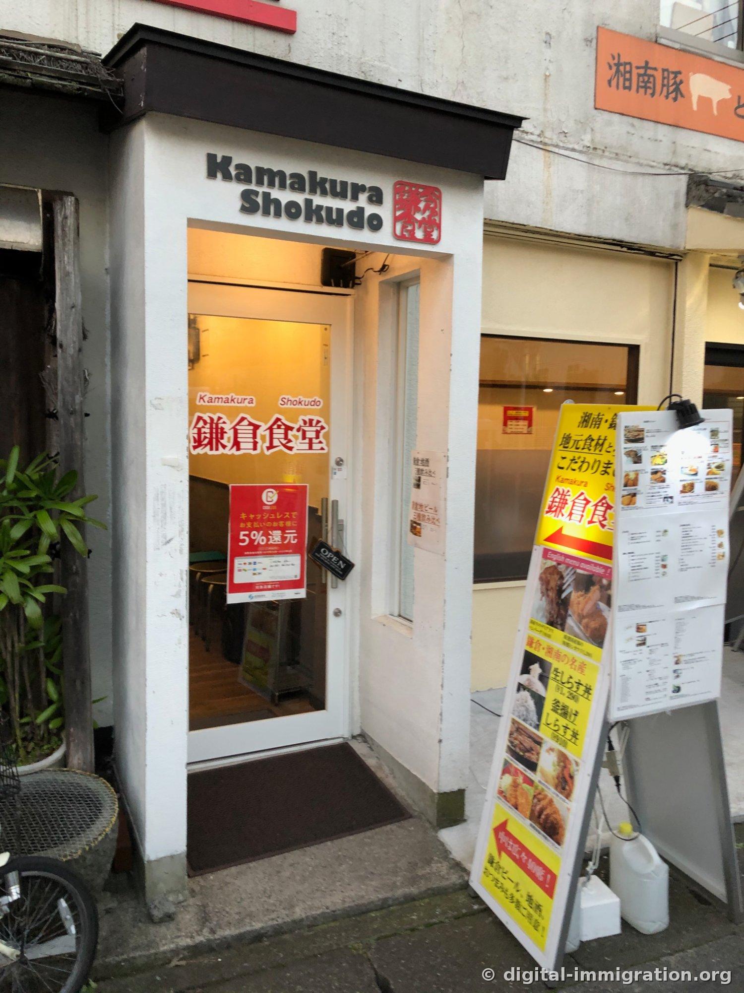 흰 벽에 Kamakura Shokudo와 鎌倉食堂 간판이 붙은 좁은 입구, 옆에 사진이 실린 노란 입간판과 영문 메뉴판이 세워져 있다
