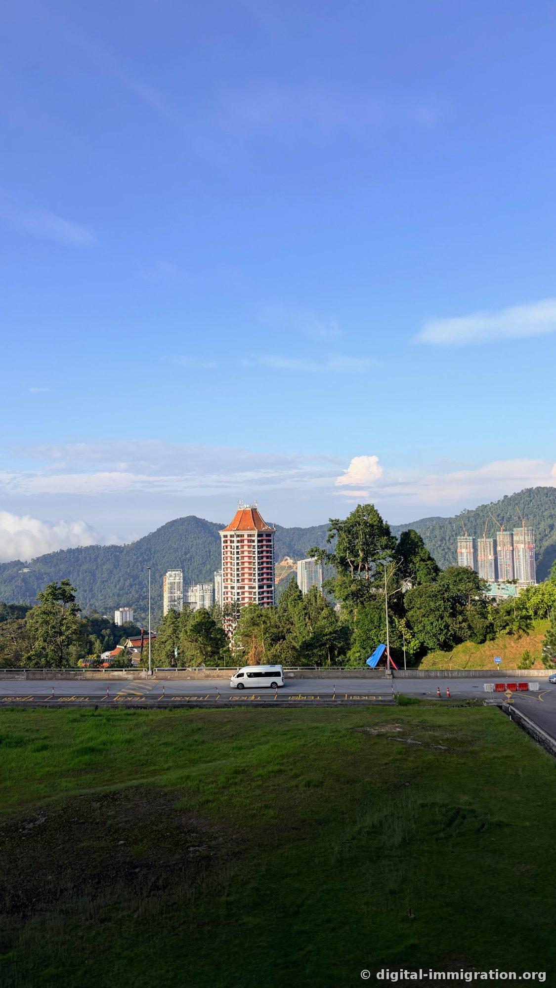 Tower-shaped hotel with orange roof and construction high-rise building under a blue sky, with taxi waiting lane and lawn in front
