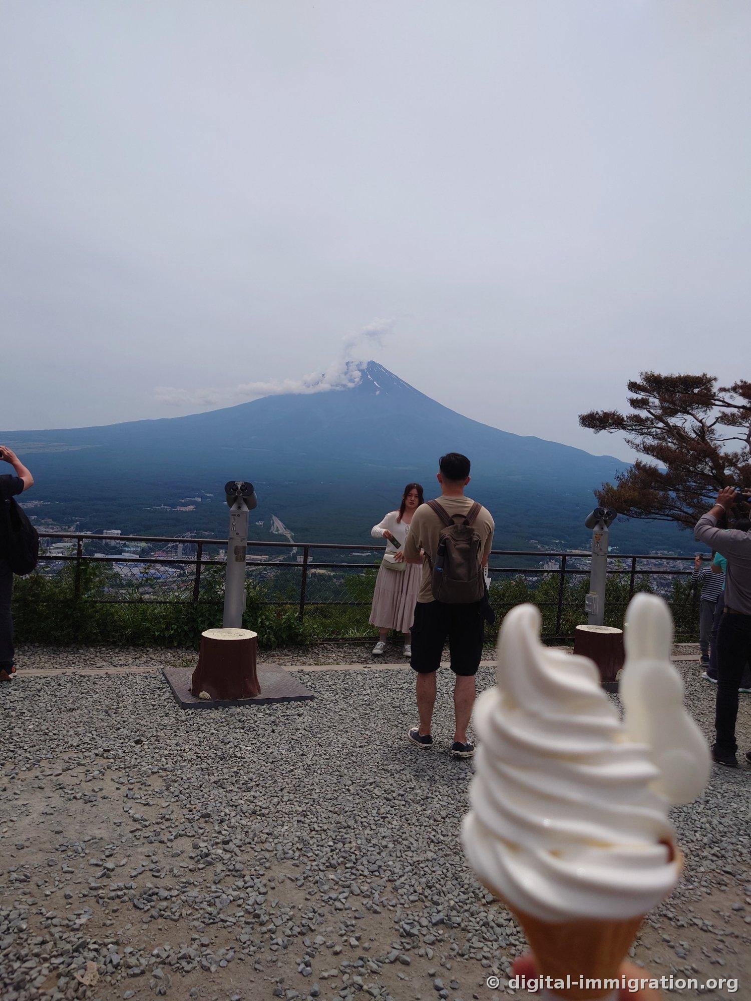 Mount Fuji with clouds, the observation deck telescopes, and people taking photos visible beyond a soft-serve ice cream cone held in hand