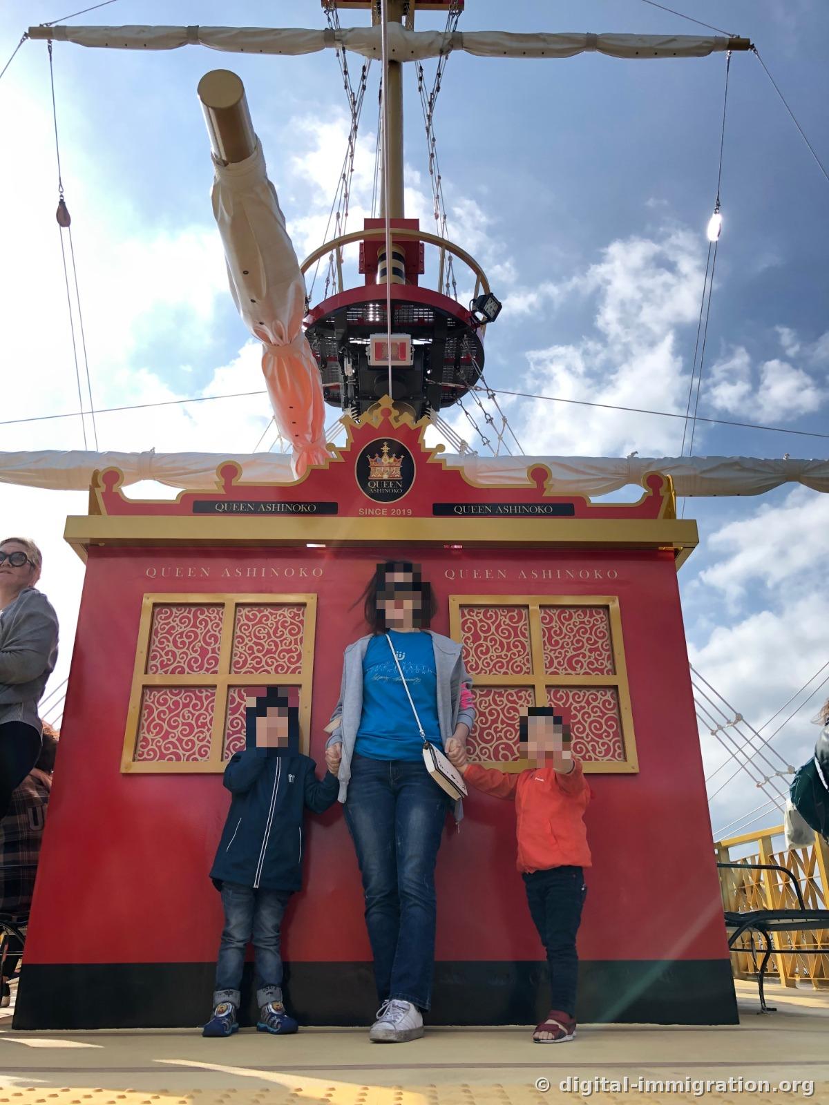 Un adulte et deux enfants debout devant le mur de la cabine rouge avec des arabesques, avec l'inscription or 'QUEEN ASHINOKO SINCE 2019' visible au-dessus et les mâts avec les voiles pliées en arrière-plan