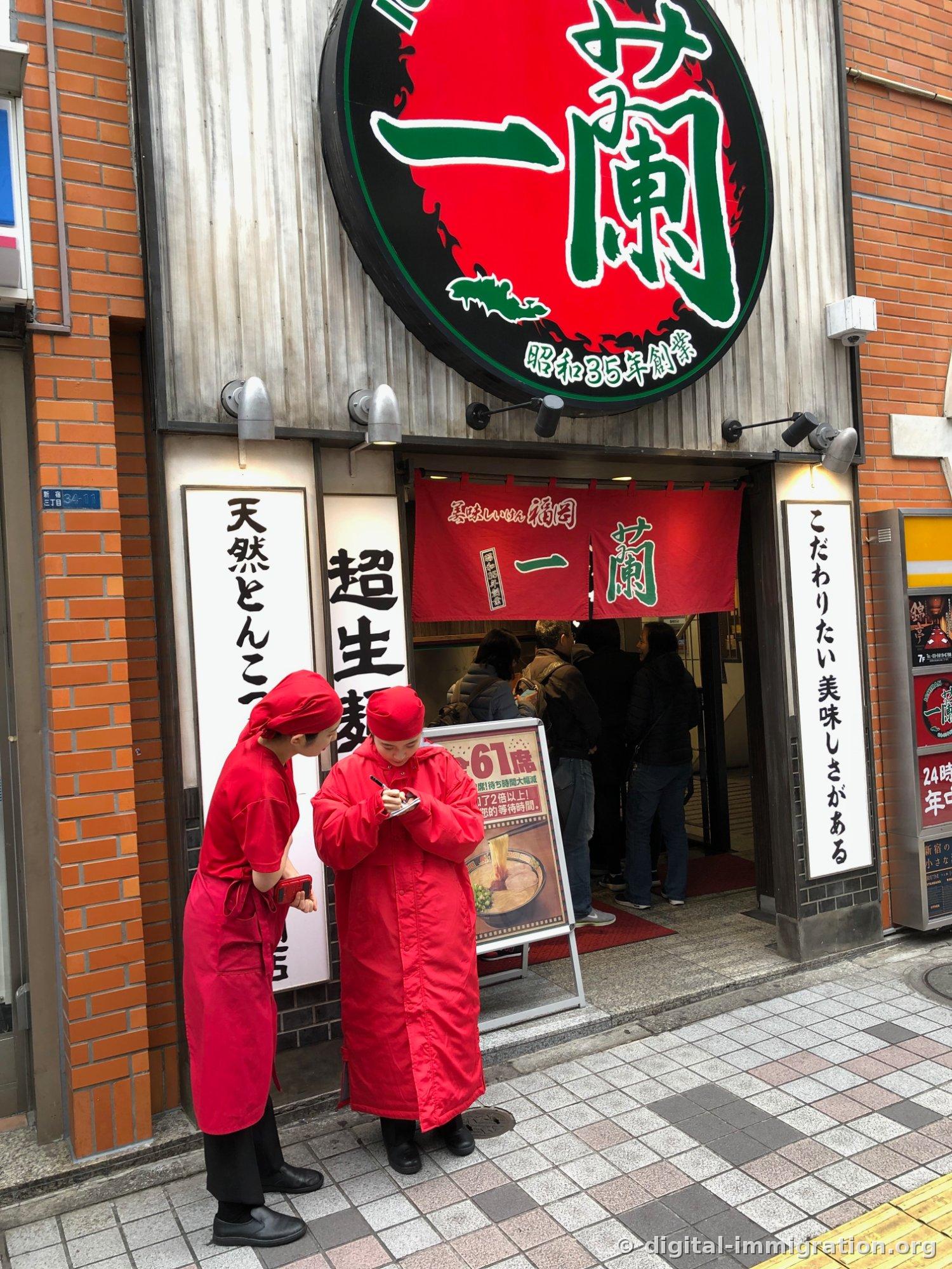 À midi, devant l'entrée de la succursale de Shinjuku 3-chome : deux employés en uniforme rouge attendent avec le registre des clients à l'entrée, sous l'enseigne circulaire rouge d'Ichiran. L'adresse « Shinjuku 3-chome 34-11 » est visible sur le panneau mural.