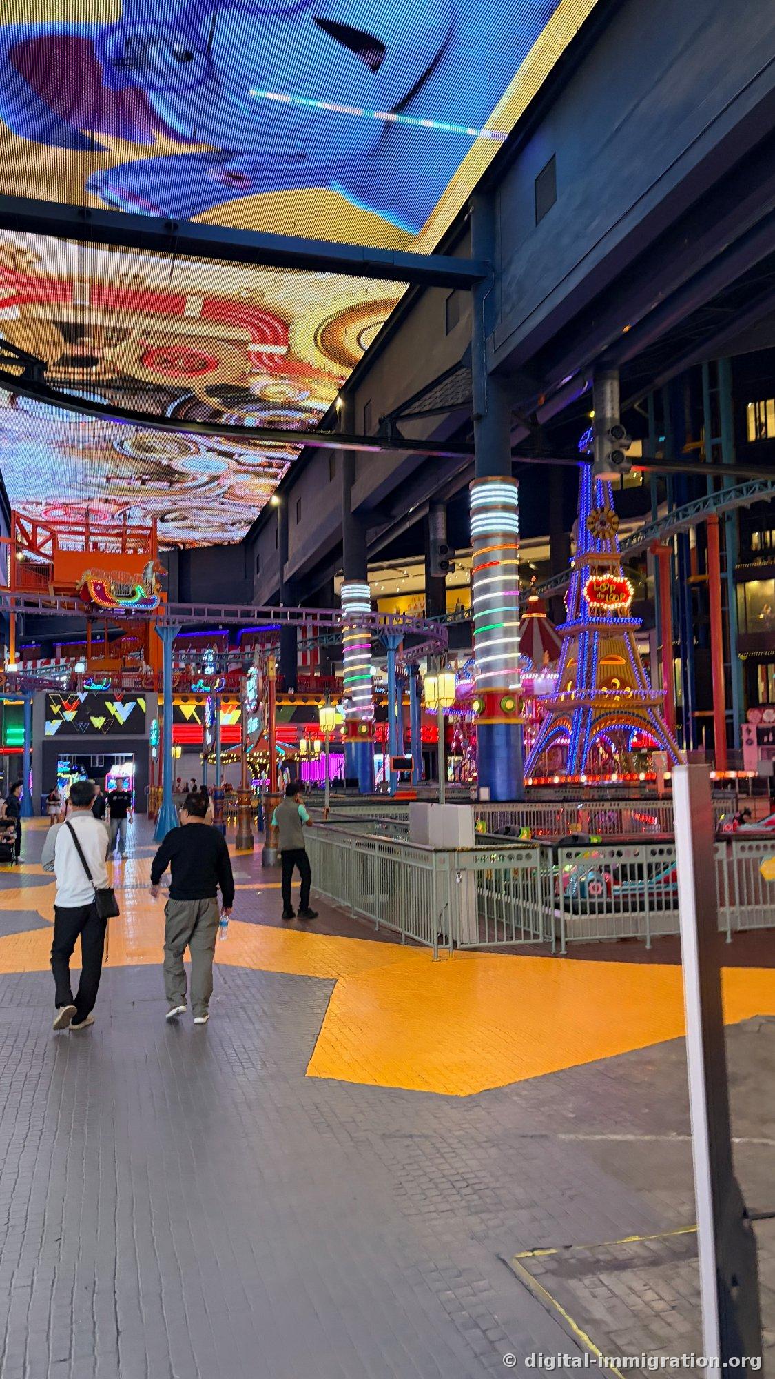 Indoor theme park corridor with yellow floor pattern and neon pillars, Loop de Loop structure on right, blue ceiling LED