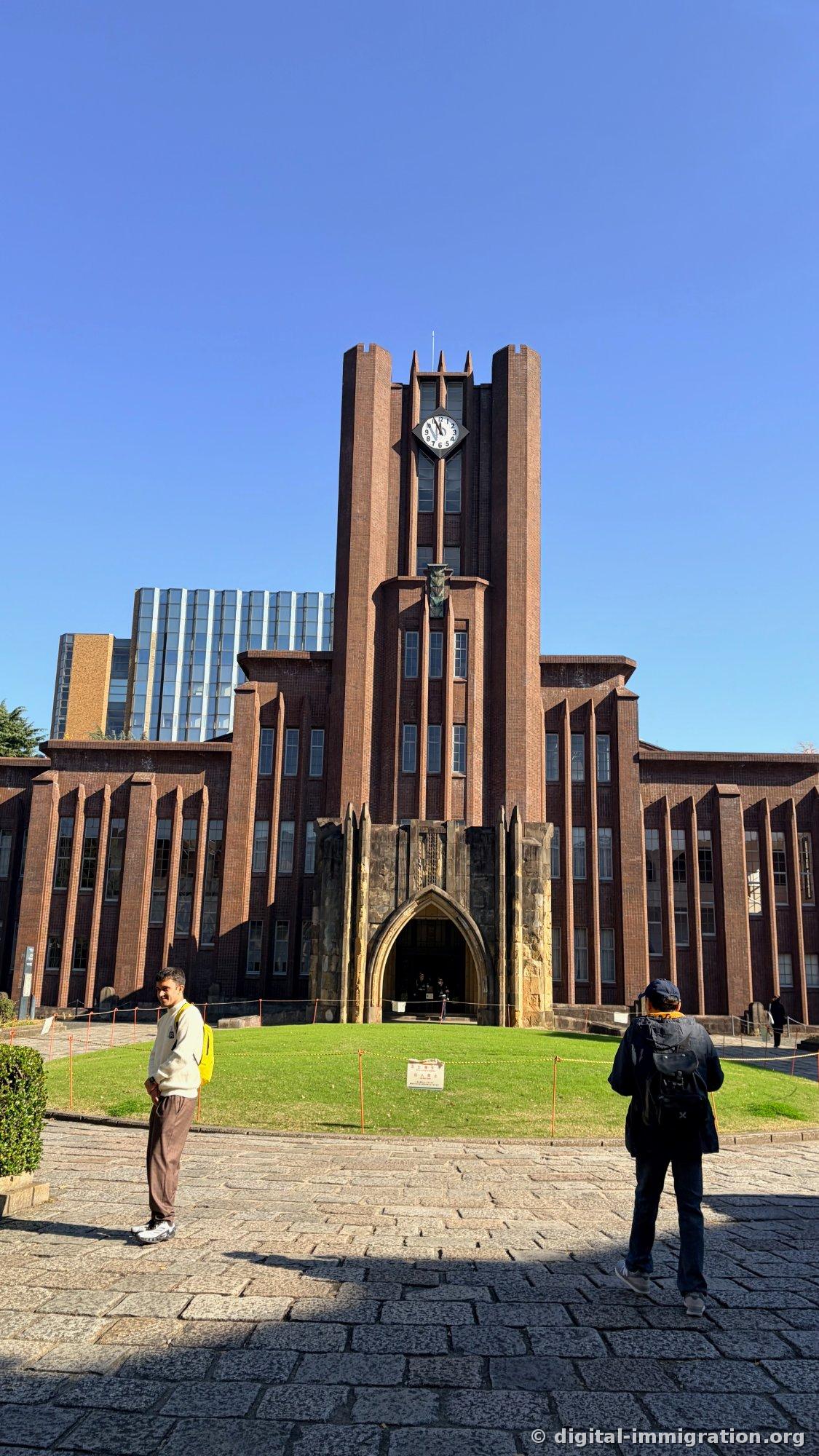 Yasuda Auditorium viewed from below on a stone-paved plaza, with a no-entry sign on the green lawn and two people gazing up at the building