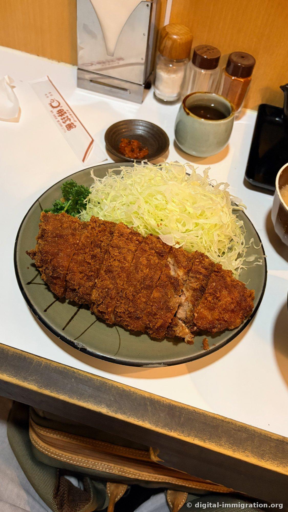 A loin katsu plate on a brightly lit white counter, featuring a large tonkatsu slice, shredded cabbage alongside, a sauce dish, red pickles, and a chopstick wrapper printed with the Tonchinjkan logo