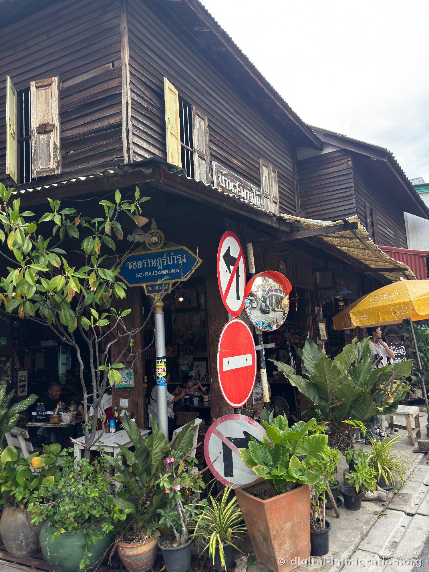 Corner wooden two-story building with SOI RAJBAMRUNG blue road sign and multiple traffic signs, diners eating at restaurant below