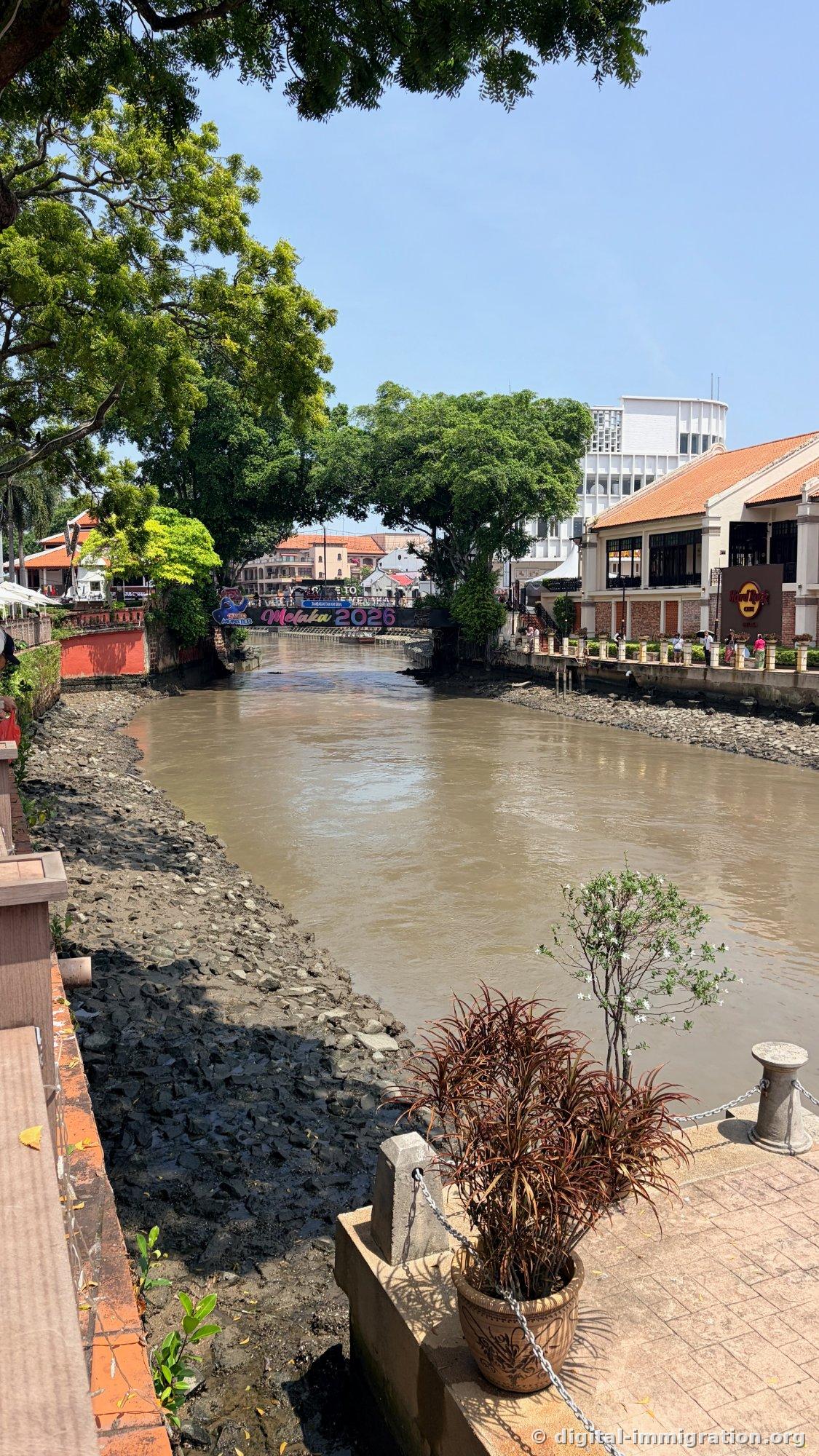 The Melaka River riverbed exposed at low tide with mudflats and pebbles visible. In the distance, a colored banner reading 'Melaka 2026' hangs on a bridge, and the Hard Rock Cafe building is visible on the right riverbank