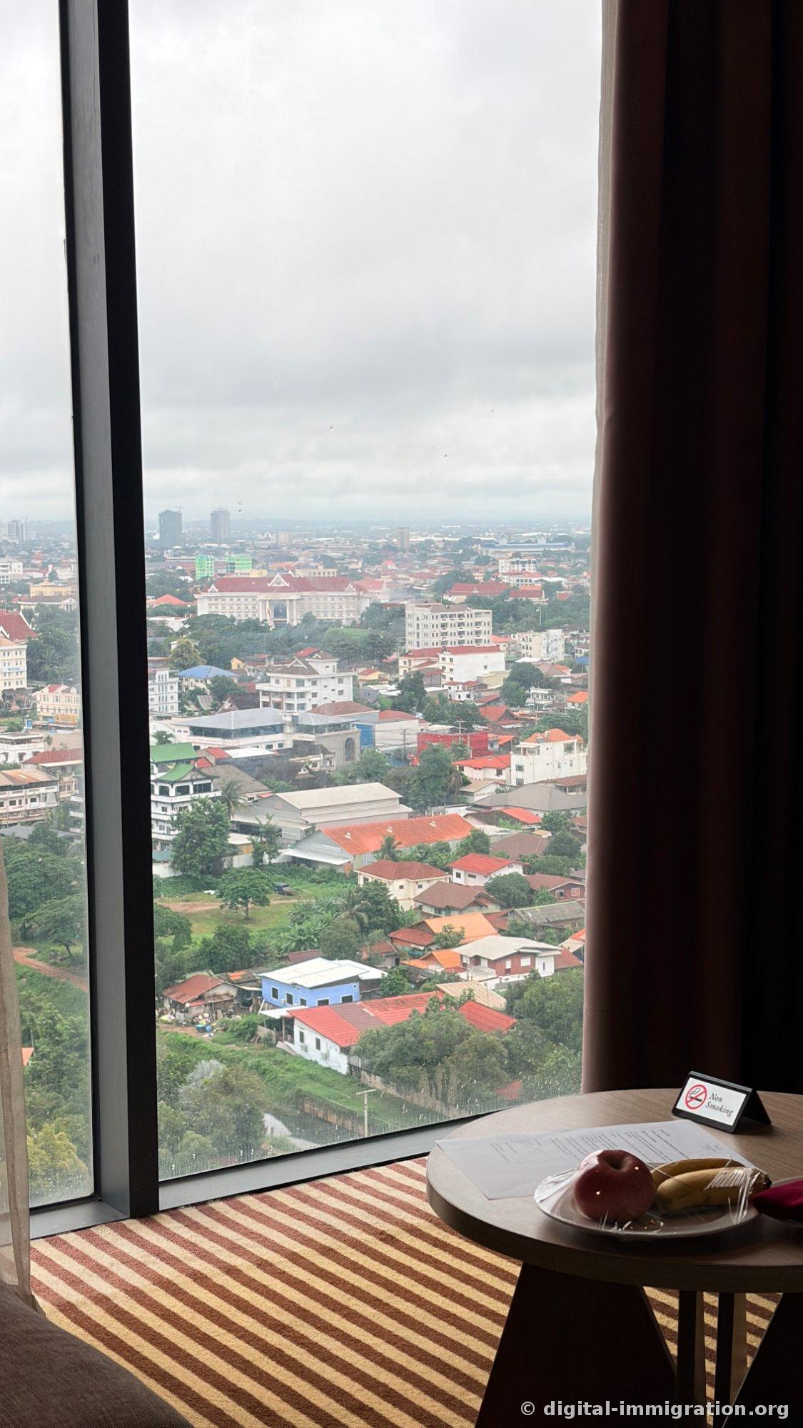 Vue depuis les baies vitrées de la chambre sur le quartier résidentiel de basse altitude de Vientiane, avec une table ronde près de la fenêtre portant une assiette de fruits de bienvenue avec pomme et banane, et un panneau « Non Smoking »