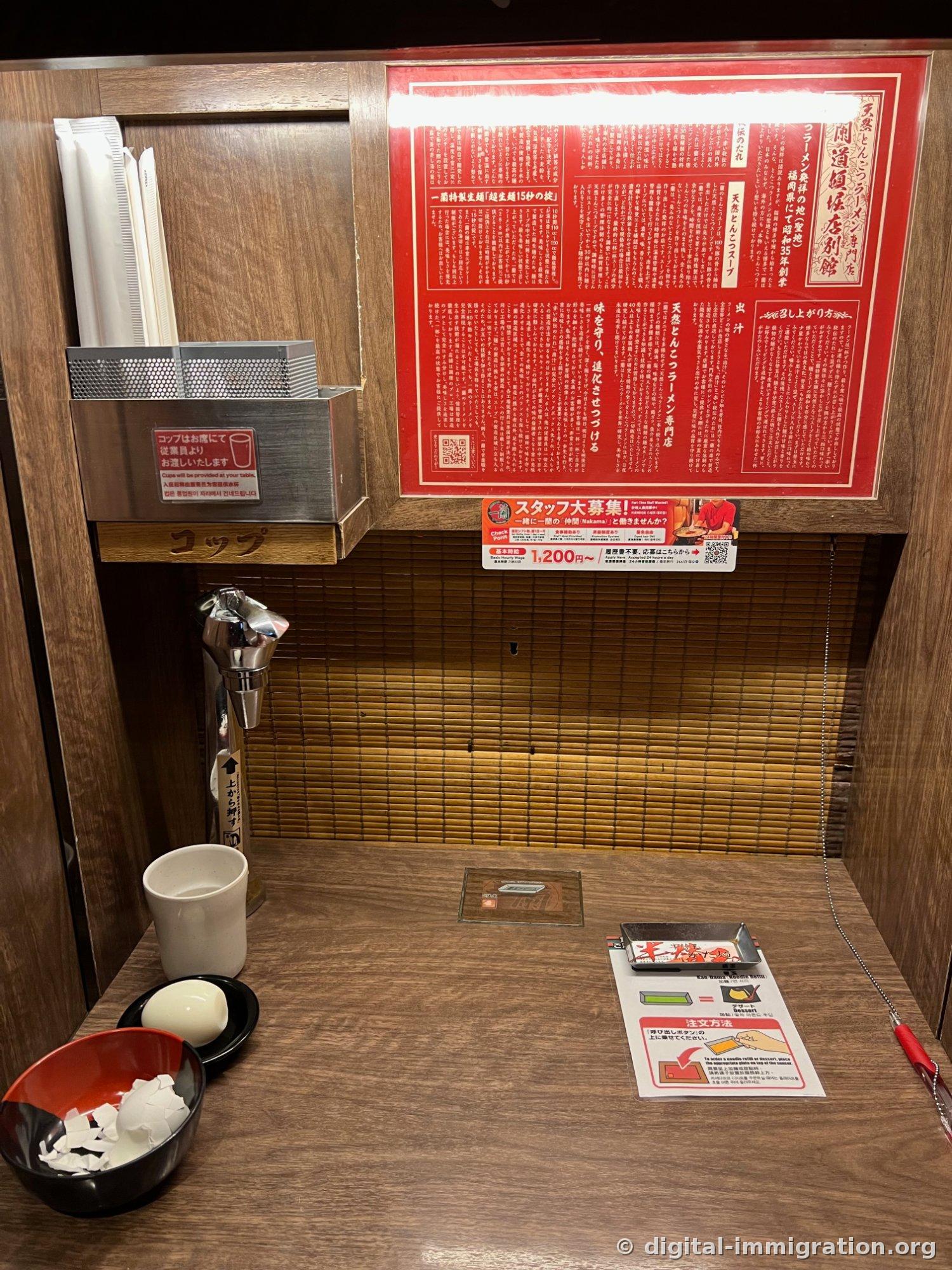 An Ichiran single-seat booth. A wooden wall with red signage, a cup storage box, a self-serve water dispenser tap, and order instruction sheet and call button on the wooden counter