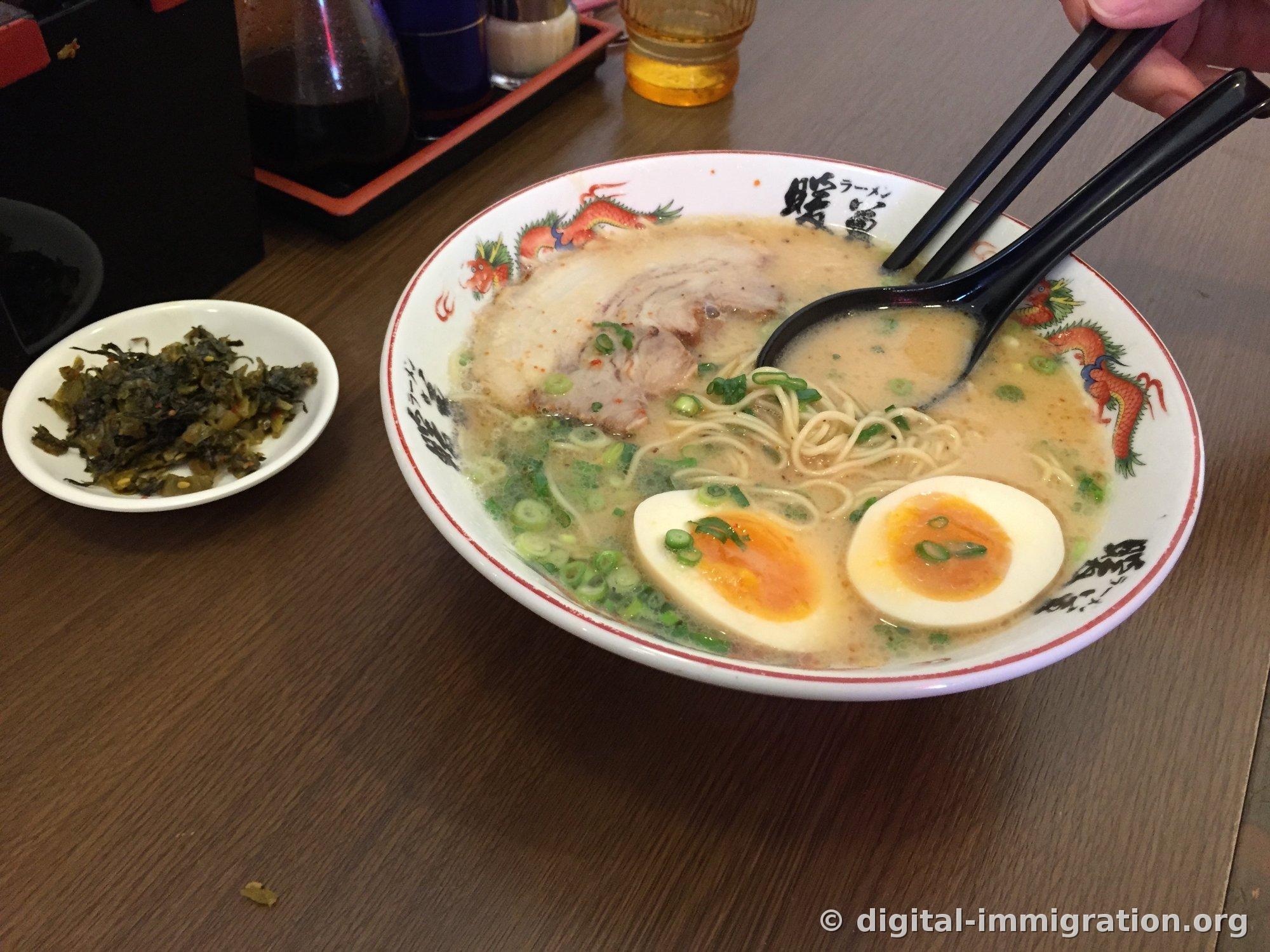 Ramen tonkotsu Tanpo servi dans un bol blanc avec motif de dragon. Deux tranches de porc rôti, deux demi-œufs mollets, oignons verts sur le dessus, une petite assiette de takana mariné à côté