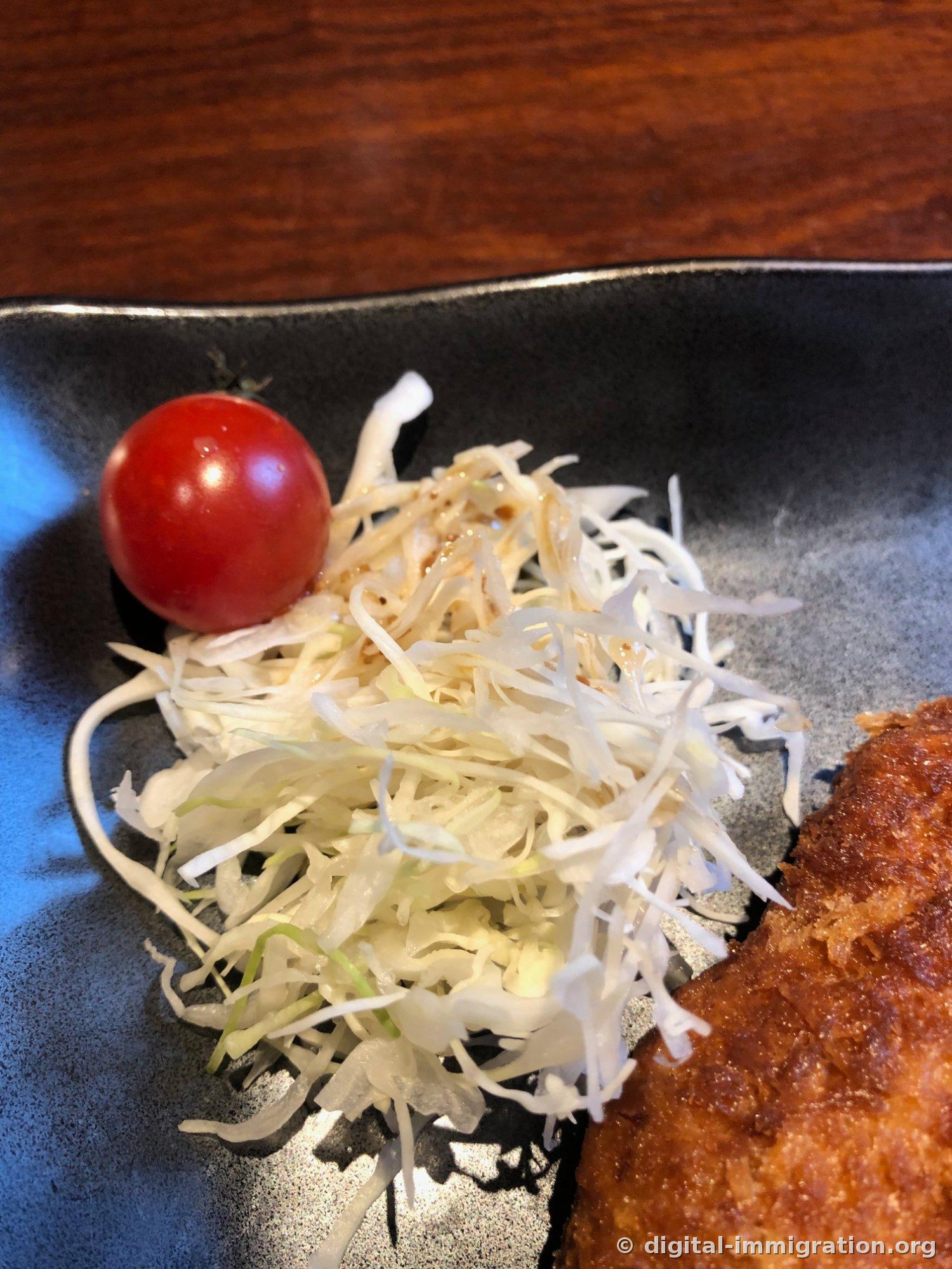 黒い皿の上に細く千切りされたキャベツの山と赤いミニトマトのクローズアップ。右下に揚げ物の一部が見える