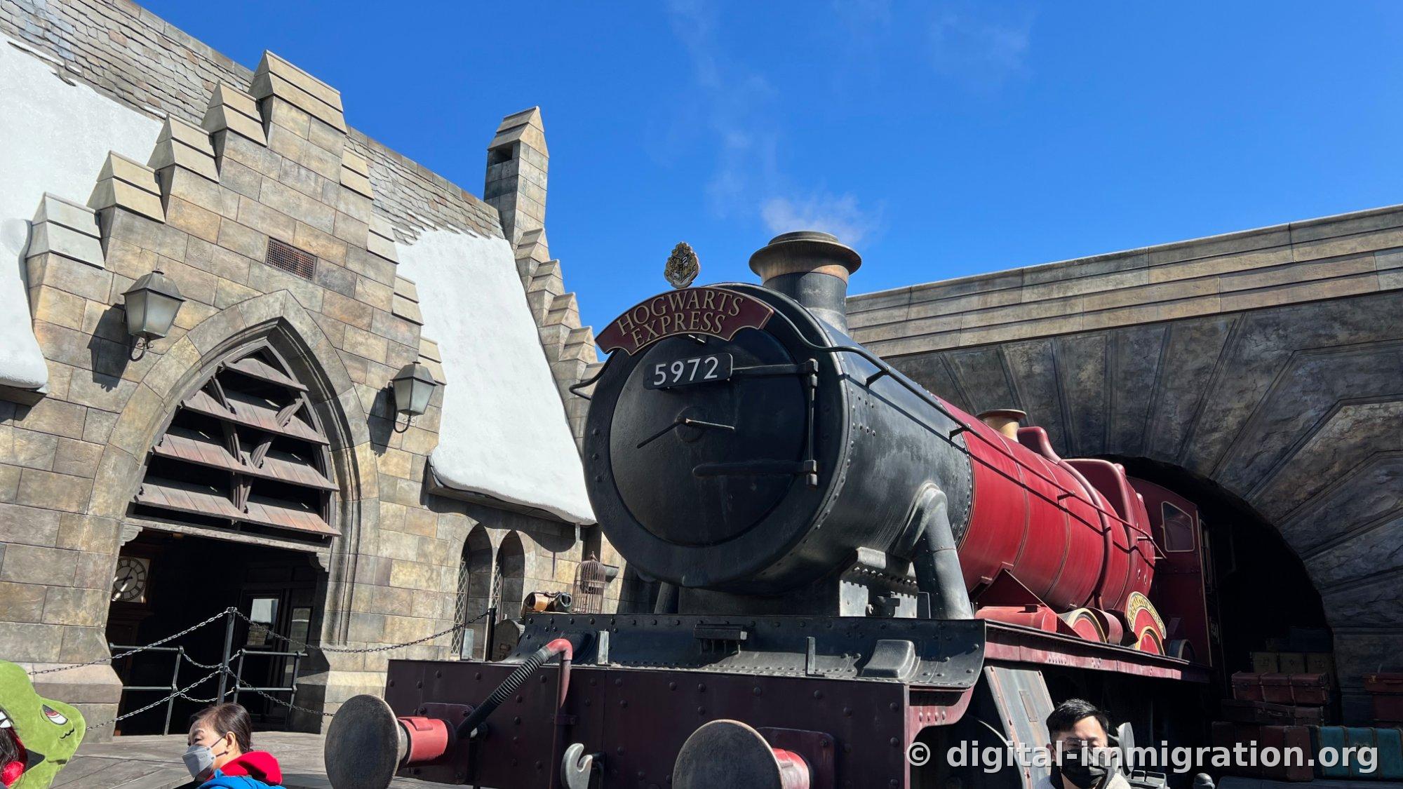 Hogwarts Express with crimson cars and black locomotive parked before Hogsmeade Station, nameplate reading HOGWARTS EXPRESS with number 5972, white steam rising from the smokestack