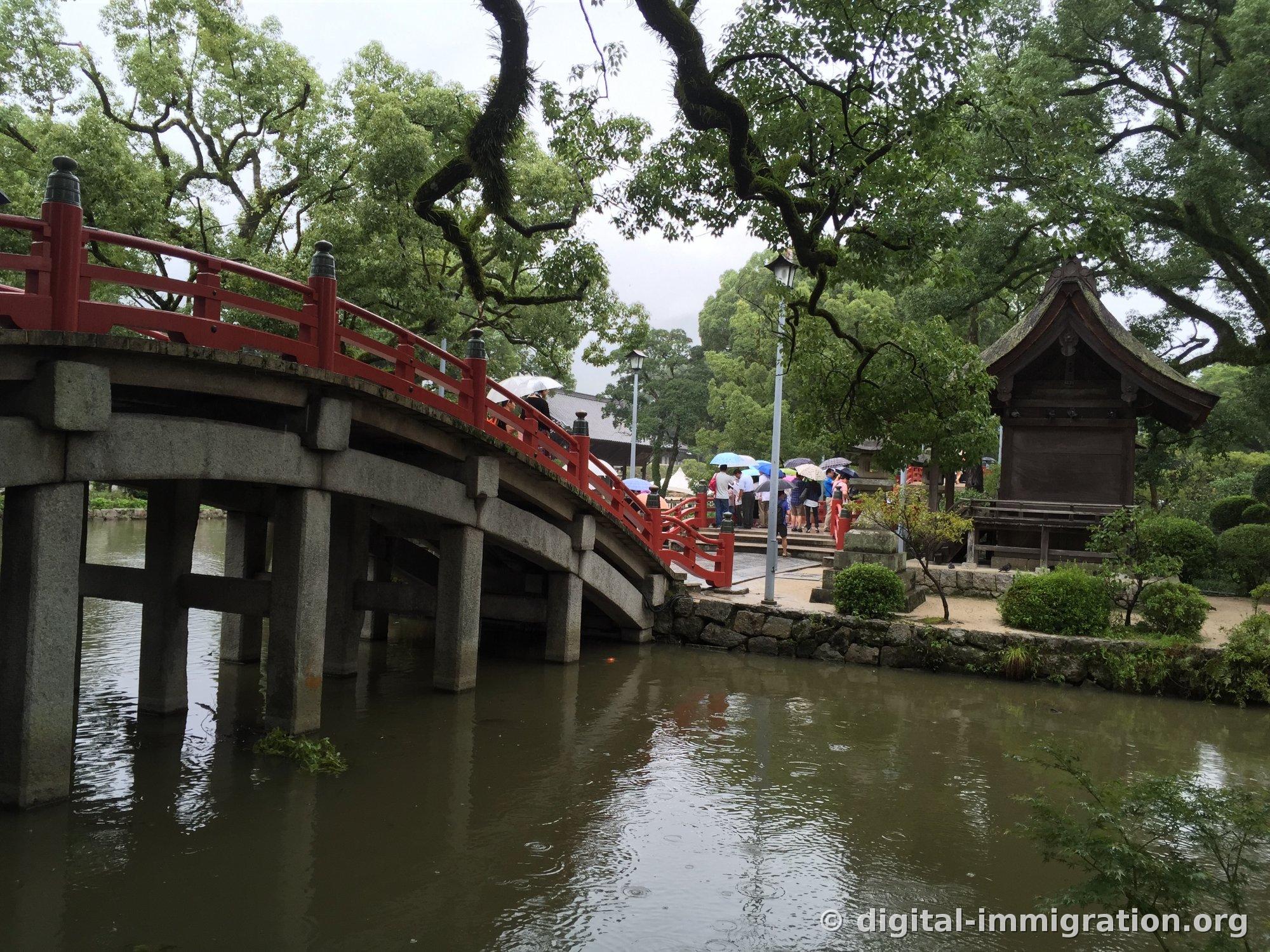 Red Daikoubashi stone bridge arch seen from the side with raindrops falling on the pond, small shrine on the right