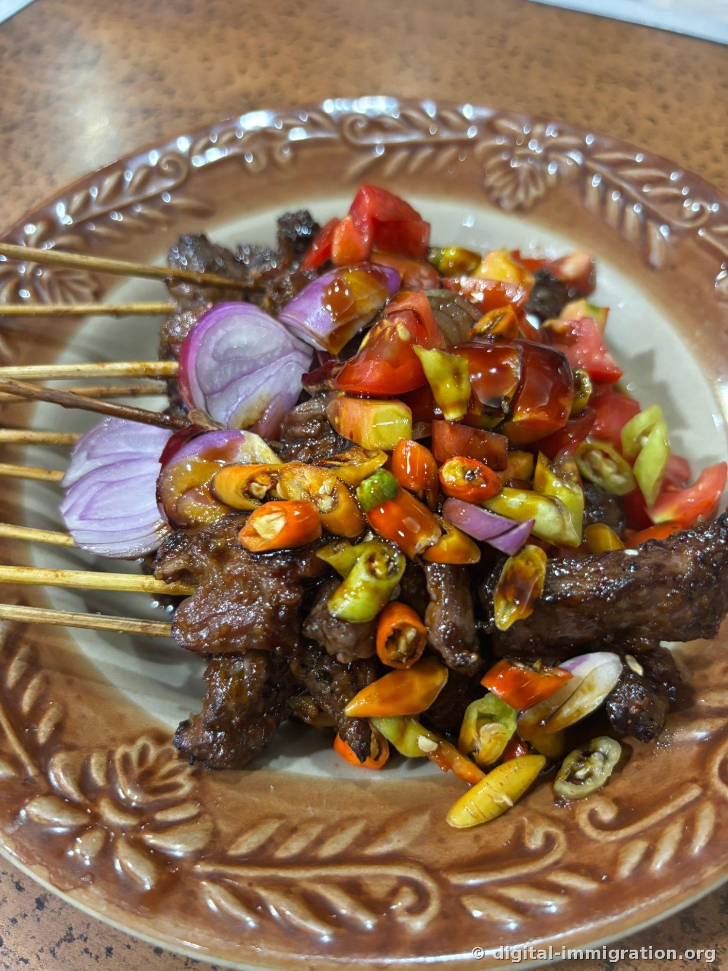 Bamboo skewers of beef satay on a patterned brown plate, heaped with fresh bird's eye chilies, tomato, and purple onion on top