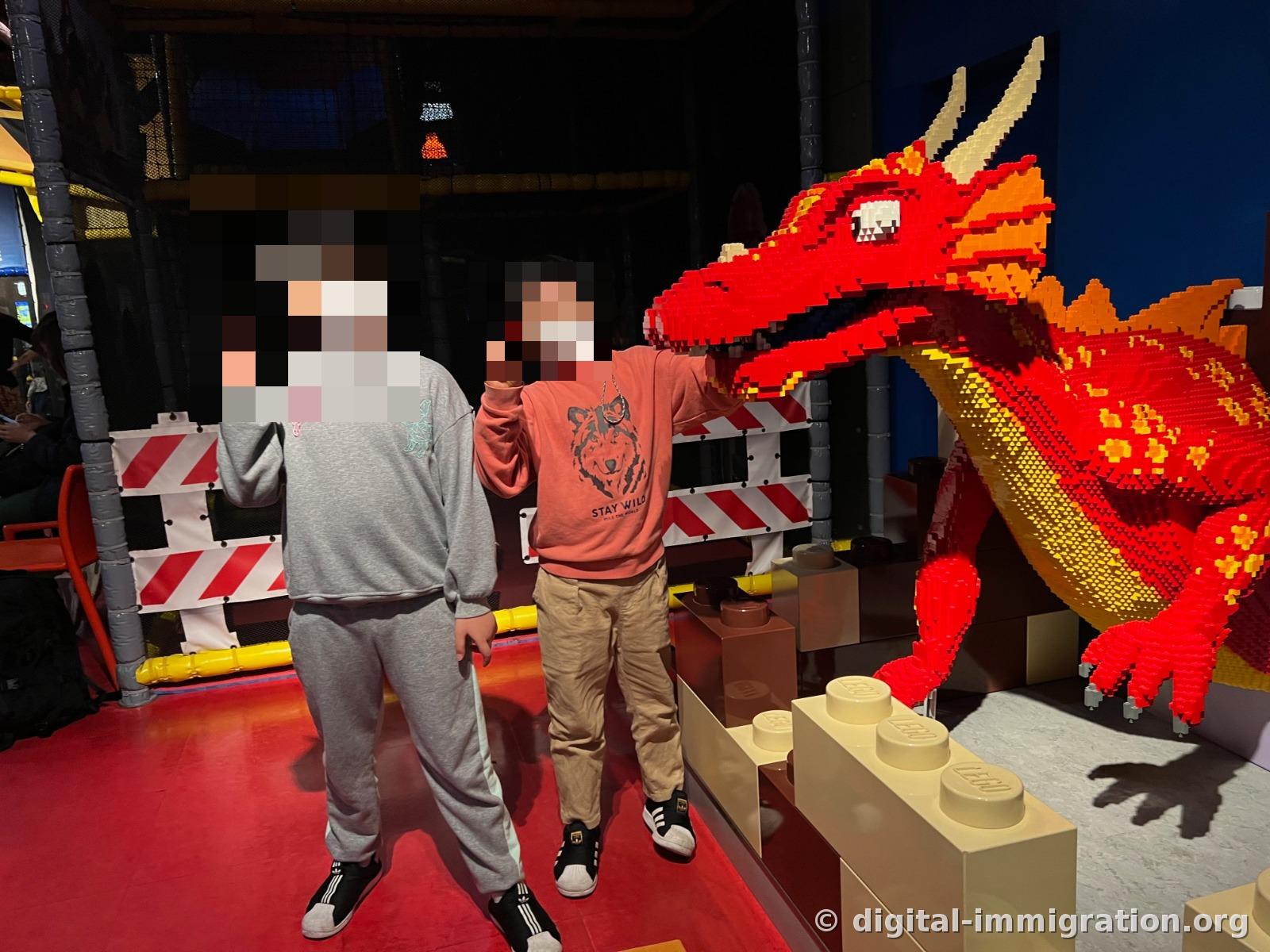 レゴブロックで作った大きな赤いドラゴンの造形物の横で子ども2人がポーズを取っており、後ろに黒いネット製の室内遊び構造物が見える
