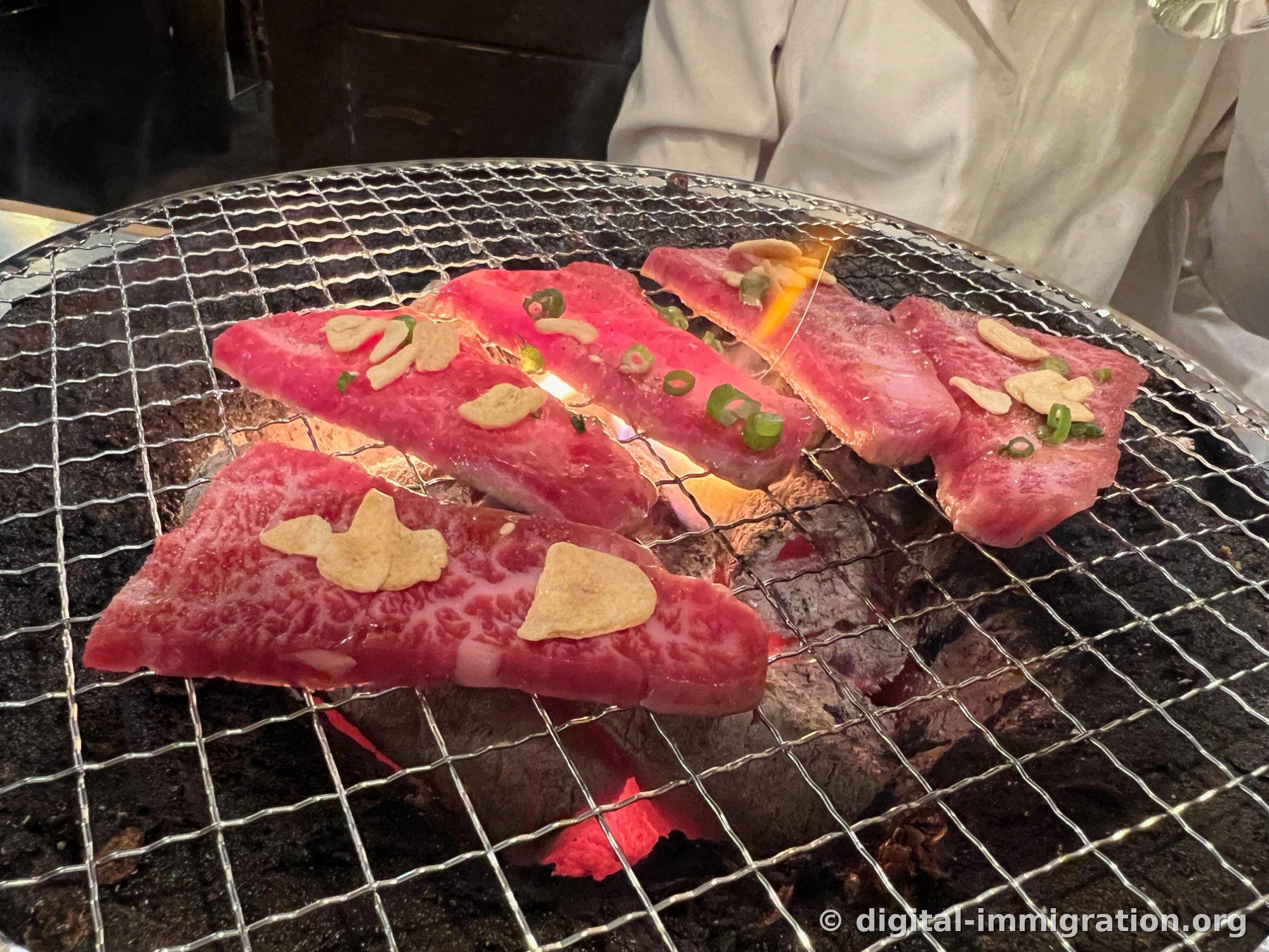 炭火網の上にマーブリングが密集した赤い牛肉が五点乗せてあって、ニンニクチップとせん切りの小ネギが乗せてある。下に炎が上がってくる