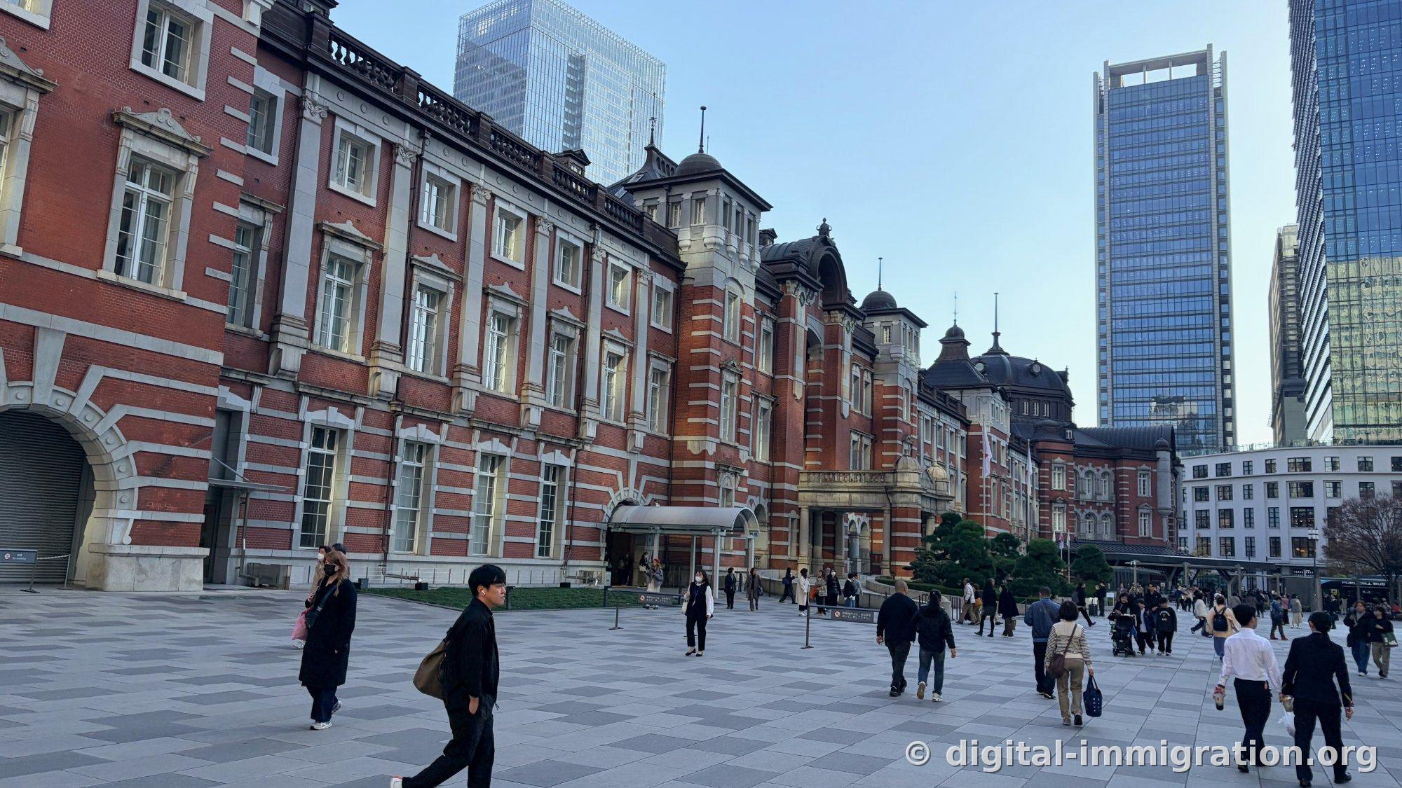 Vue en angle de la longue façade en brique rouge de la Gare de Tokyo Marunouchi depuis la place Marunouchi, avec les gratte-ciel en verre alignés derrière, et des passants traversant la place