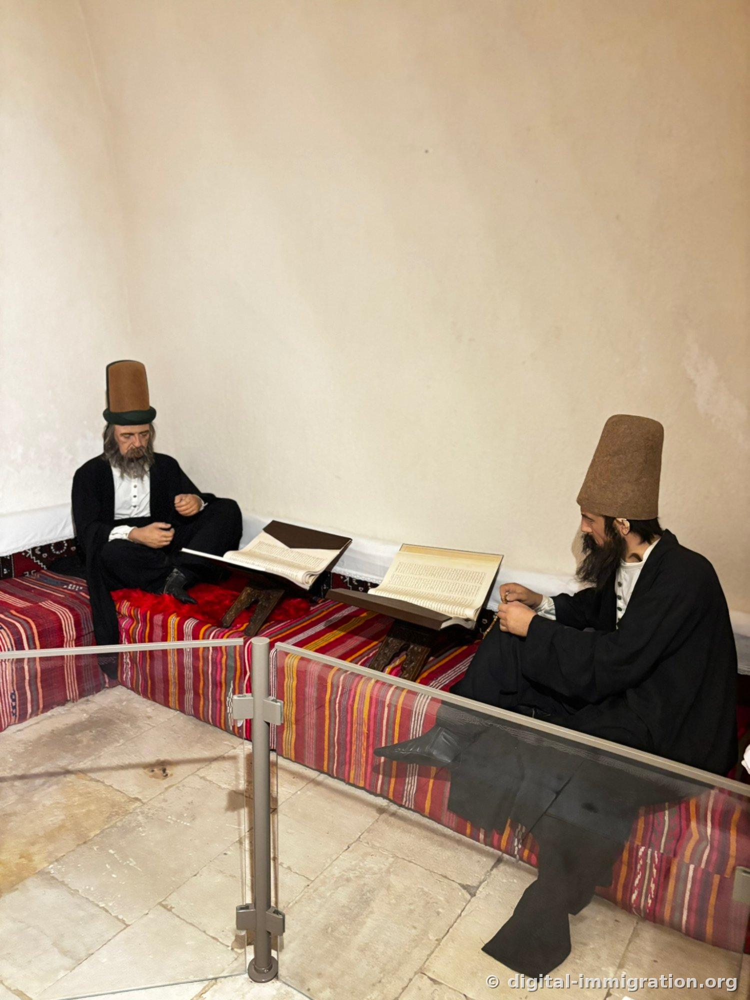 Deux figures de cire portant manteau noir et chapeau brun assis sur un piédestal kilim, lisant des livres ouverts sur un pupitre