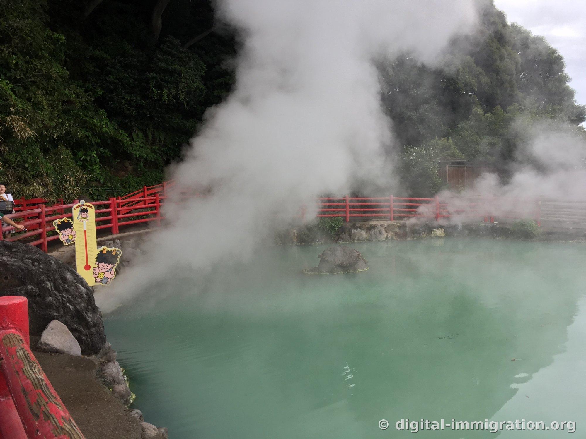 Un bassin d'eau chaude de couleur vert laiteux et une pancarte jaune de thermomètre illustrée avec un personnage de démon, avec de la vapeur s'élevant densément au-dessus de l'eau