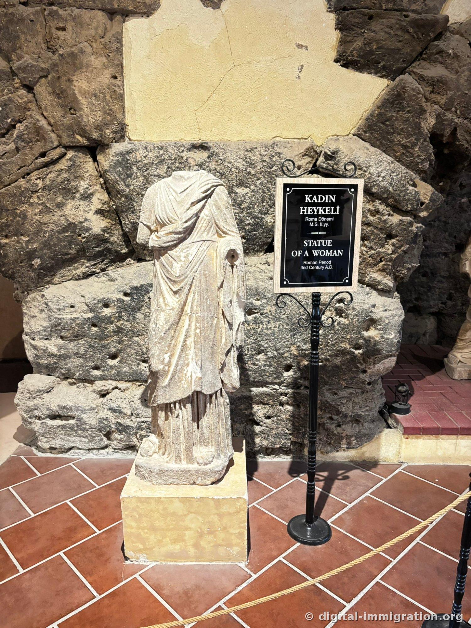 Headless and armless female statue standing on a yellow pedestal, placard reading 'KADIN HEYKELI / STATUE OF A WOMAN' placed beside it