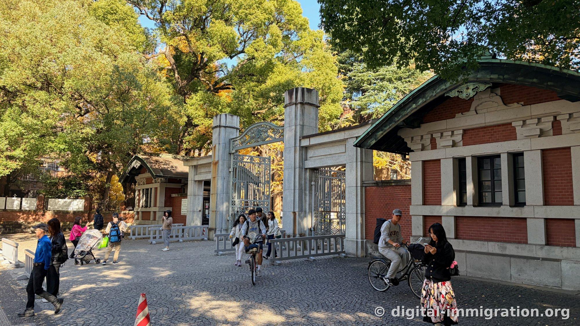 Red brick gatehouse with green copper roof and iron decorative gate, stone-paved ground in fan pattern, a student on a bicycle and a family with a stroller passing by