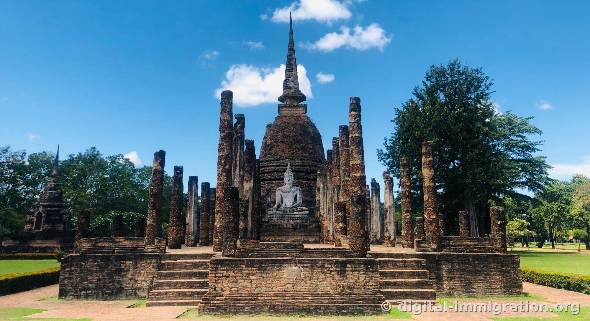 The bell-shaped brick spire of Wat Sa Si and a white seated Buddha in front, with thick stone columns stretching left and right and fluffy clouds against a blue sky