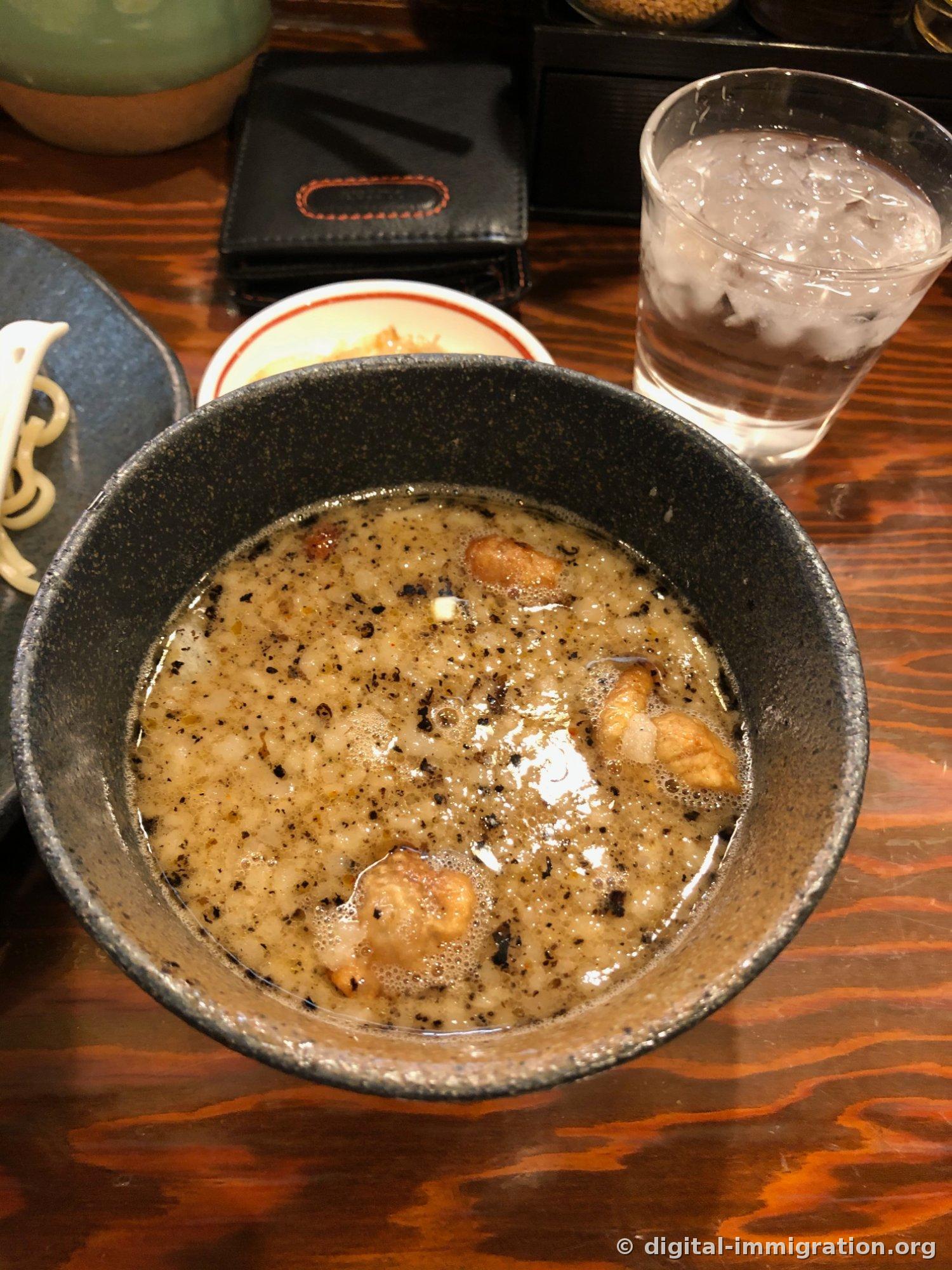Le bouillon tsukumen dans un bol en céramique noire. Un bouillon trouble et épais avec des particules noires de shoyu brûlée flottant à la surface et des morceaux de motsu brûlés visibles