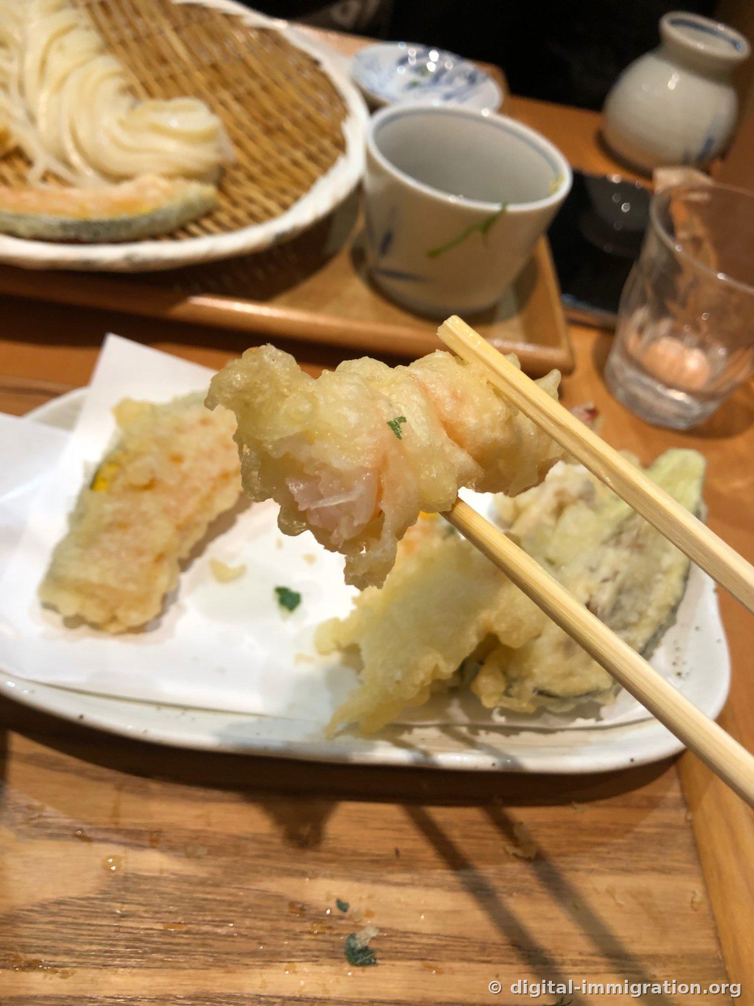 箸で持った薄い衣のエビ揚げ、皿にはかぼちゃとナス揚げが残っていて奥に陶製の酒瓶と湯呑が置かれている