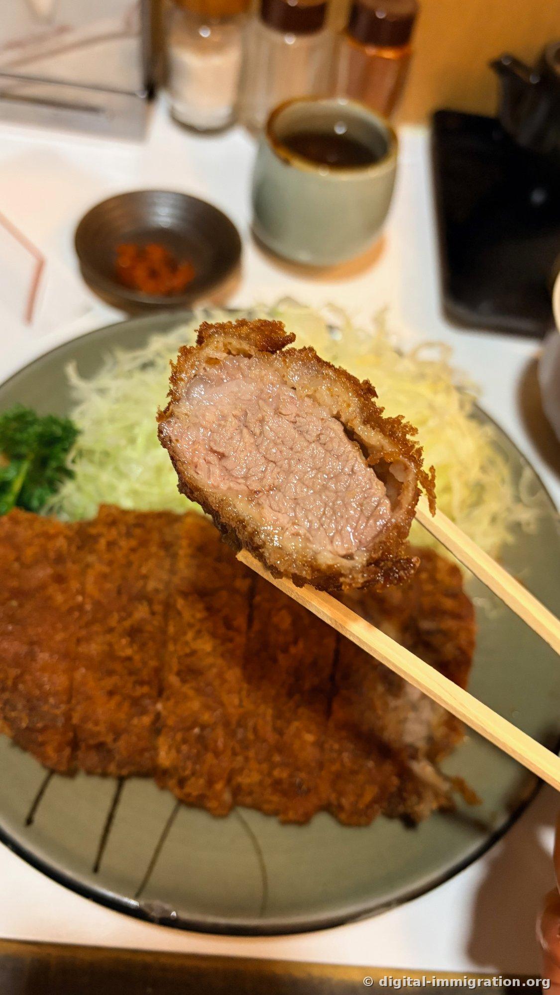 Cross-section of a loin katsu piece held with chopsticks, showing light pink-tinged meat with tight grain inside the breading