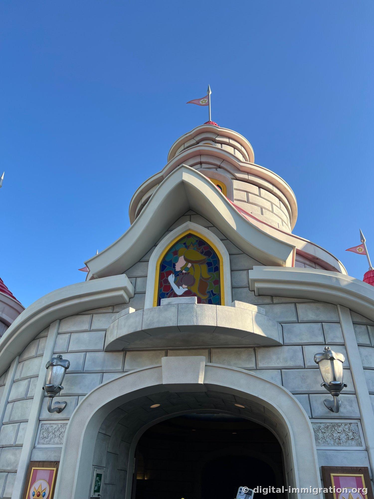 Peach's Castle exterior wall viewed from below. A pink-roofed spire, star-patterned flags, and a stained glass window with Princess Peach are visible