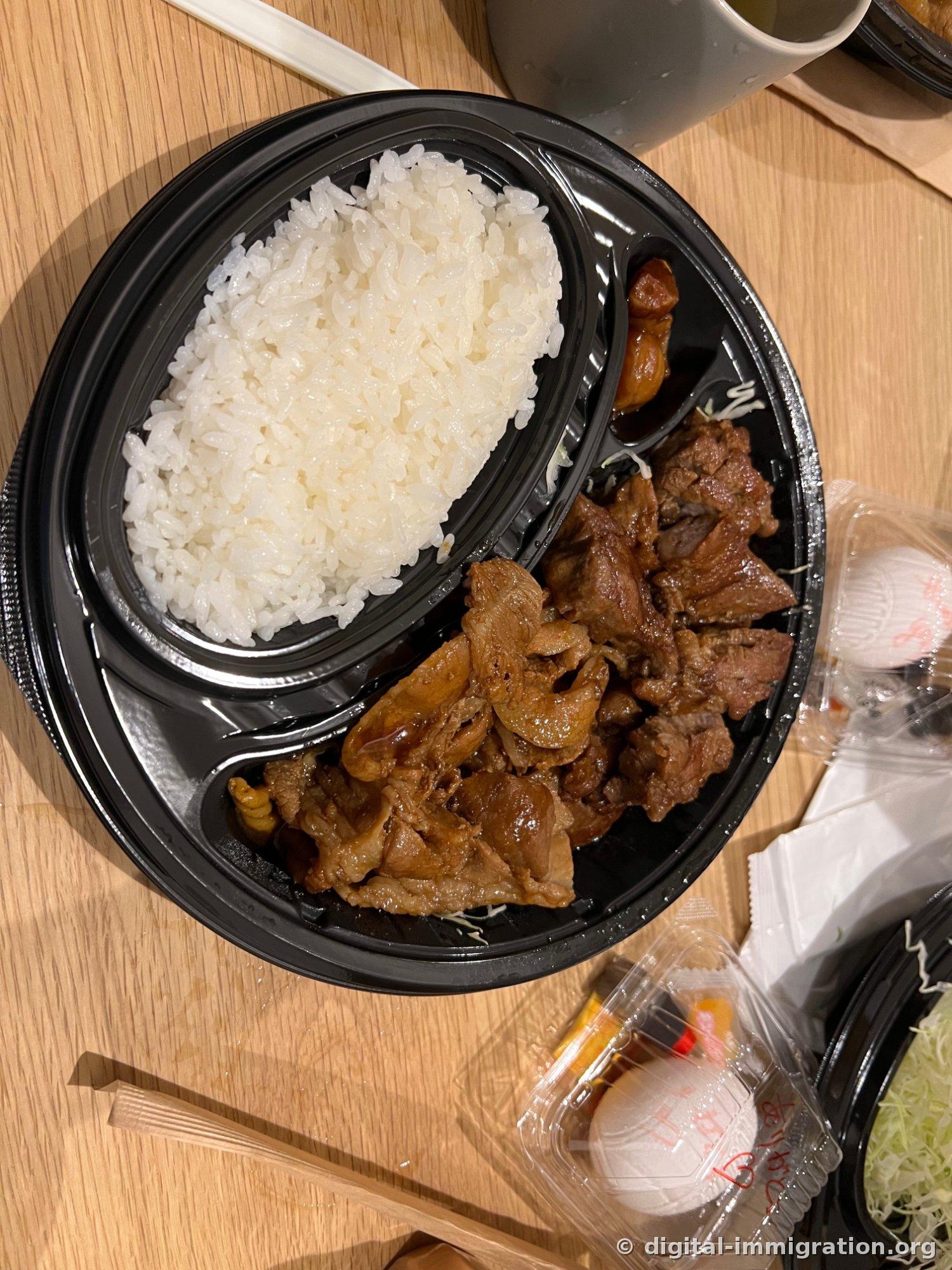 A takeout bento box set on a wooden table. A black round container with white rice on one side and soy-sauce-glazed pork on the other, accompanied by a small plastic container with onsen tamago and sauce, wooden chopsticks, and a soup cup.