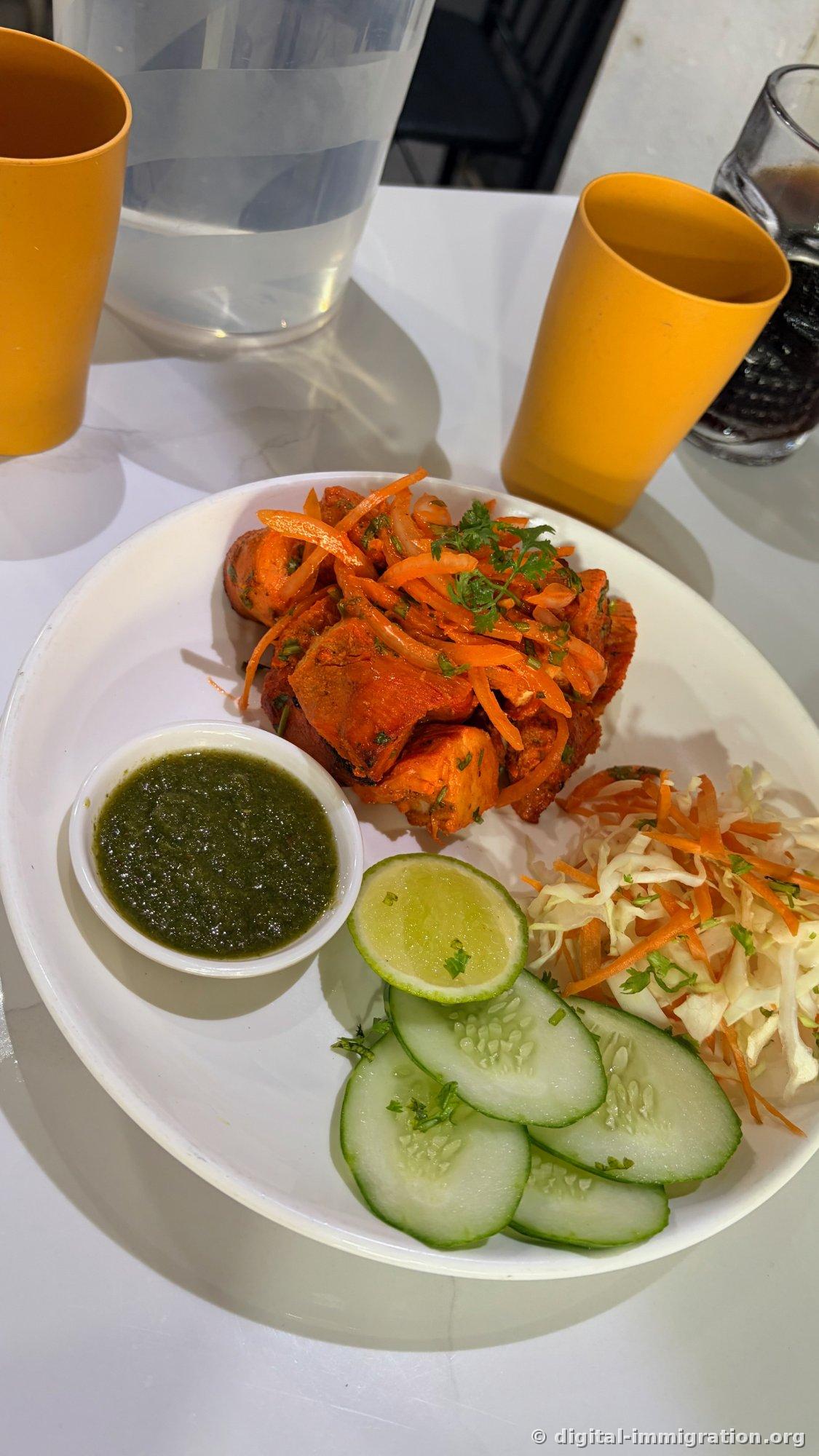 Poulet tandoori rouge sur une assiette blanche avec oignons et carottes hachés assaisonnés, coupelle de chutney à la menthe verte, tranche de concombre et salade de chou