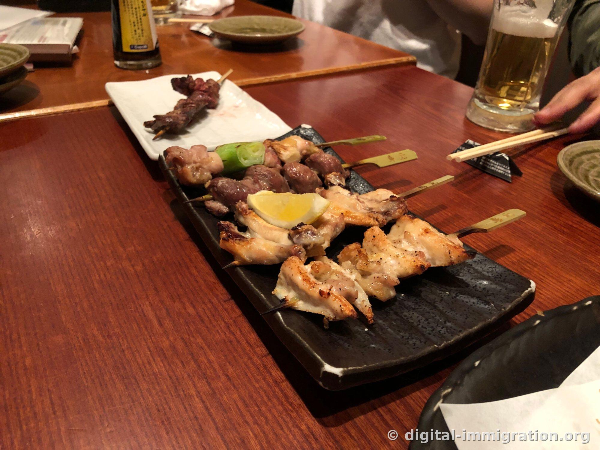 木炭火烤的鸡肉串特写，盛在黑色盘子里，右侧是拿着啤酒杯的手和筷子架