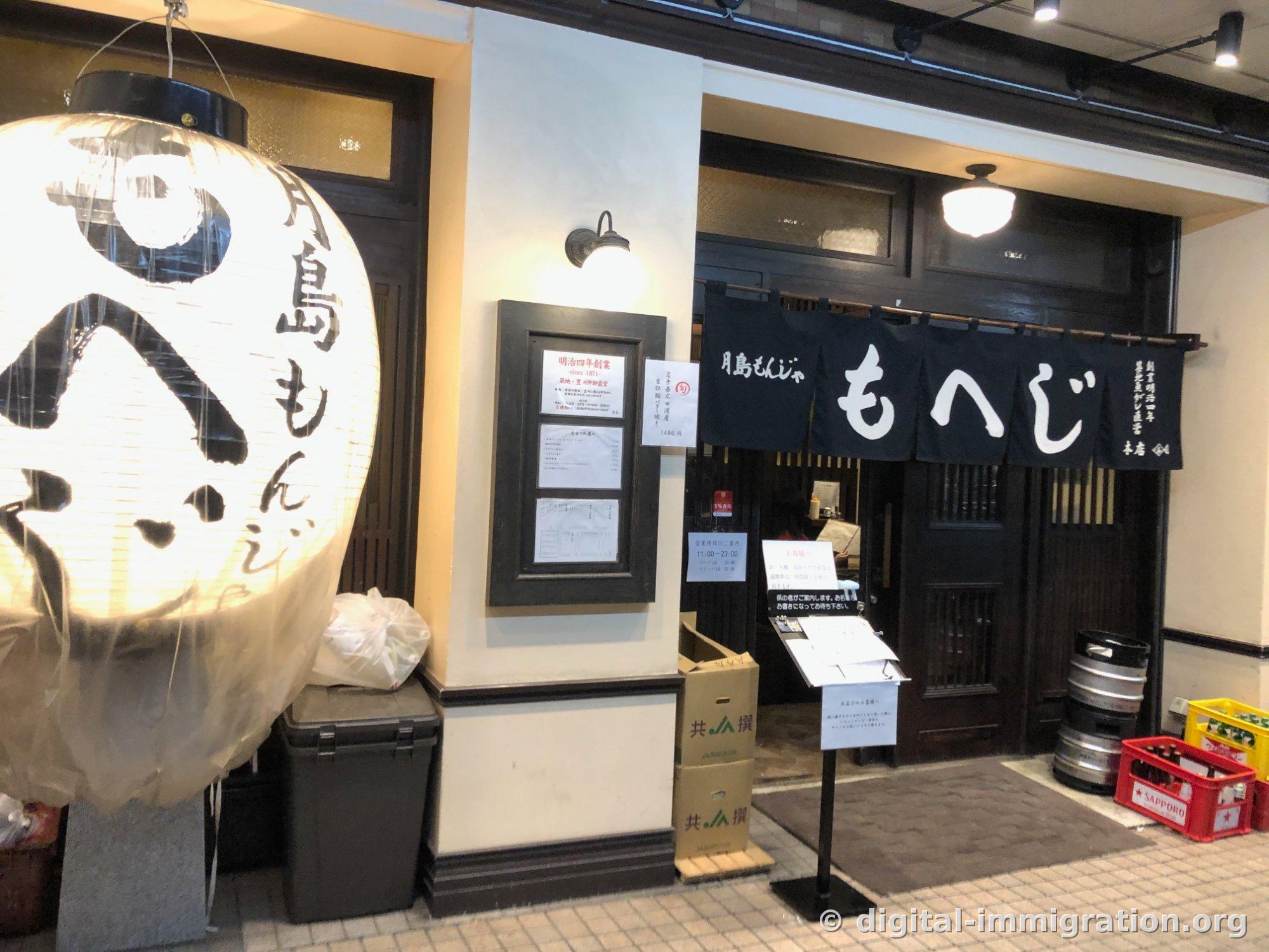 '月島もんじゃ もへじ 本店' 남색 노렌이 걸린 목조 입구와 커다란 흰 초롱, 옆에 사포로 맥주 박스