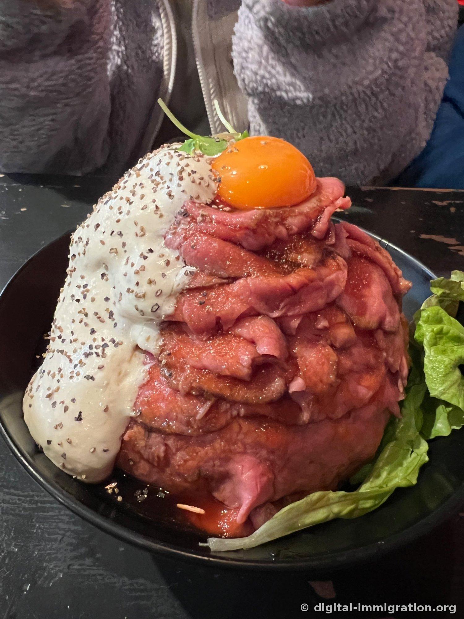 Gros plan d'un roast beef donburi servi dans un bol noir, avec du roast beef rosé empilé en montagne, un jaune d'œuf cru au sommet et une sauce blanche parsemée de poivre qui dégouline