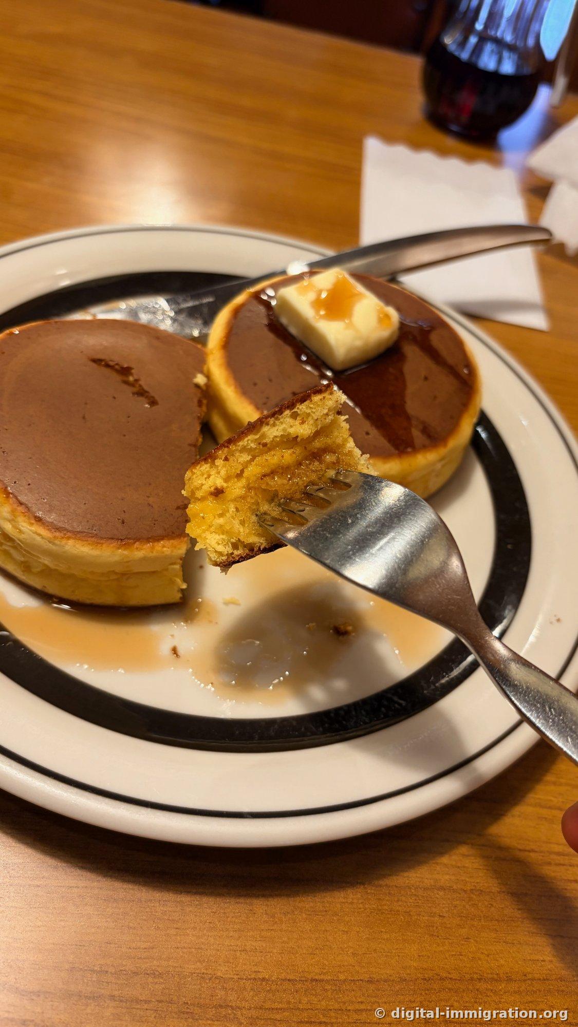 A cut piece of pancake held with a fork, showing the golden interior and syrup-soaked cross-section, with a knife resting beside the plate