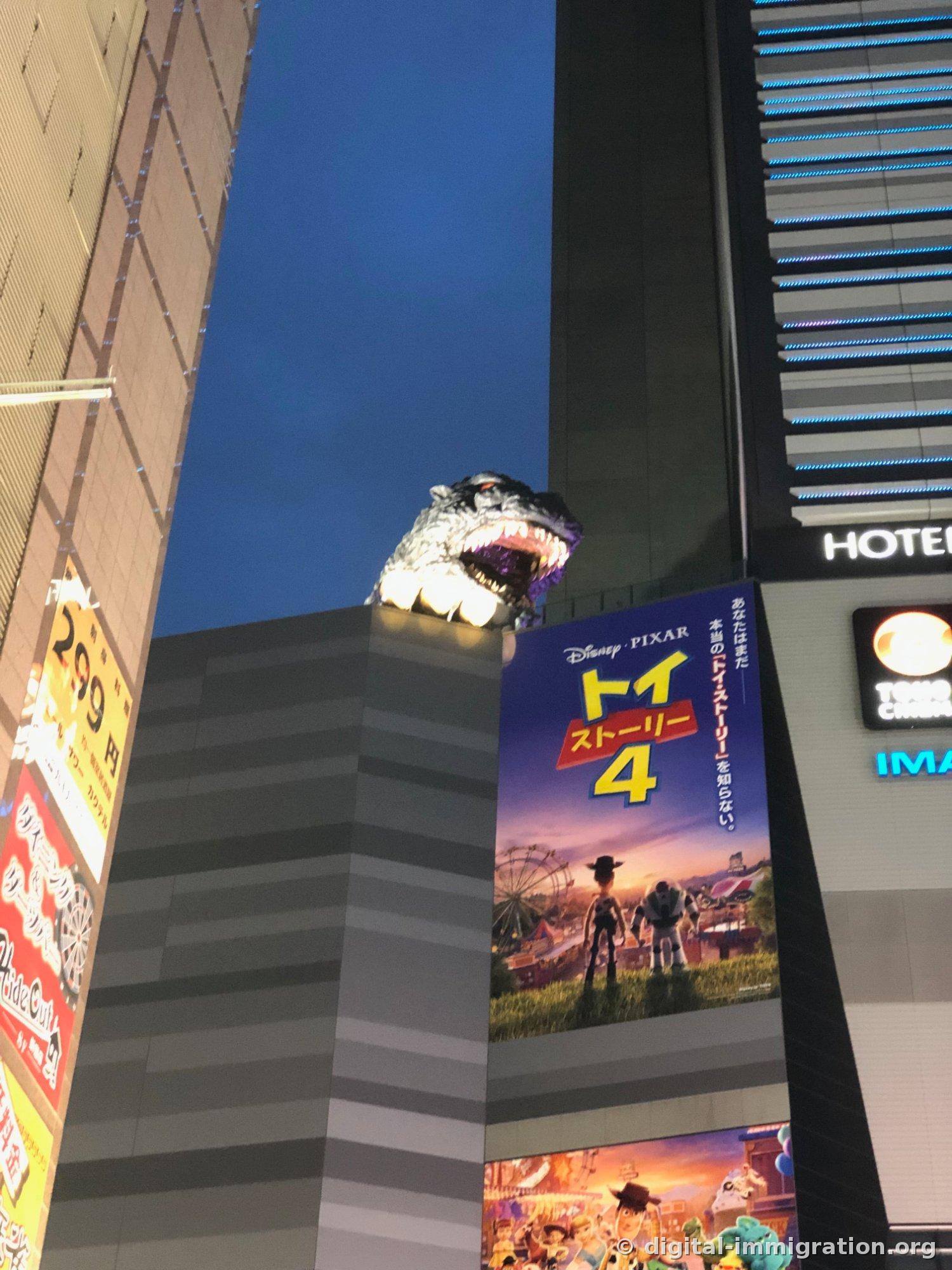 La tête de Godzilla sur le toit du bâtiment sous un ciel bleu au coucher du soleil, avec la grande affiche Toy Story 4 en dessous, et l'enseigne HOTEL à droite