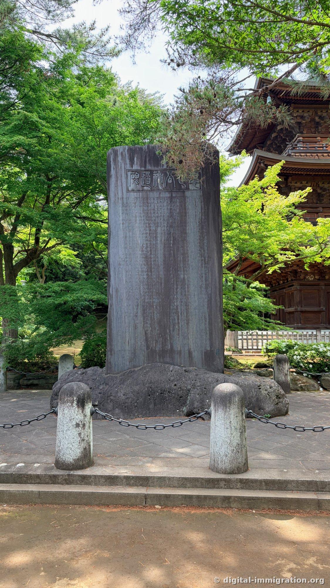 A black stone monument standing on a base surrounded by iron chains, with the eaves of a wooden three-tiered pagoda visible behind it