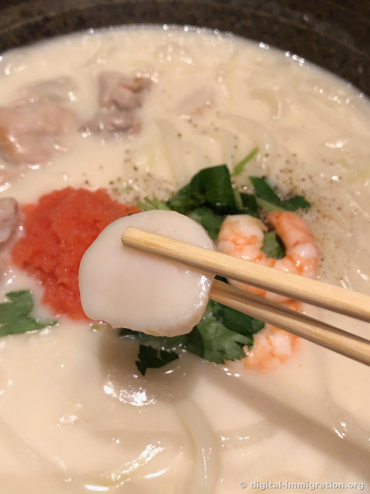 A thick scallop lifted with chopsticks, with shrimp and mitsuba visible in the background