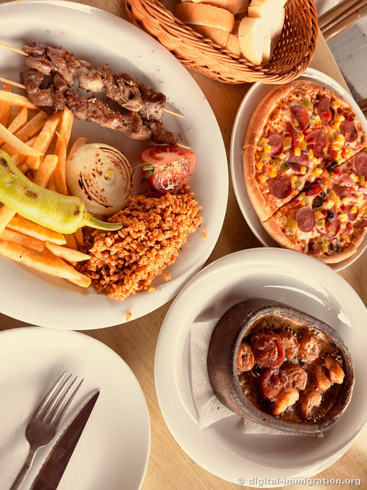 A wooden table holding plates of grilled kebab skewers, french fries, bulgur pilaf, a basket of bread, a pizza with sucuk, and a bubbling clay pot of stew