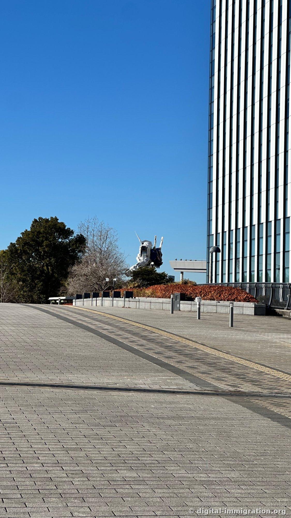 A wide paved walking path ending with the Unicorn Gundam's head and shoulders visible above trees and brown winter shrubs, a glass curtain wall high-rise building on the right