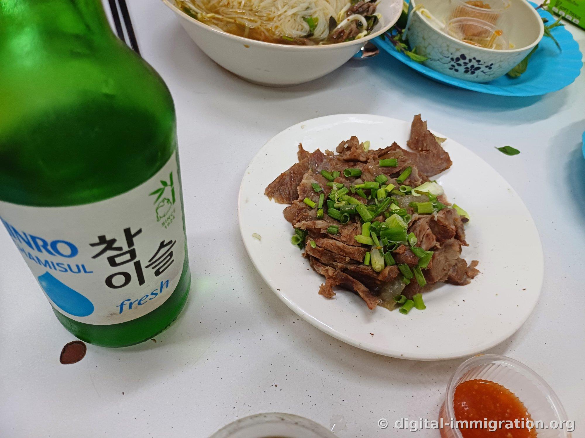 餐桌上参岛烧酒瓶旁边放着撒葱段的水煮牛肉盘，后面是米线碗和辣椒酱碟