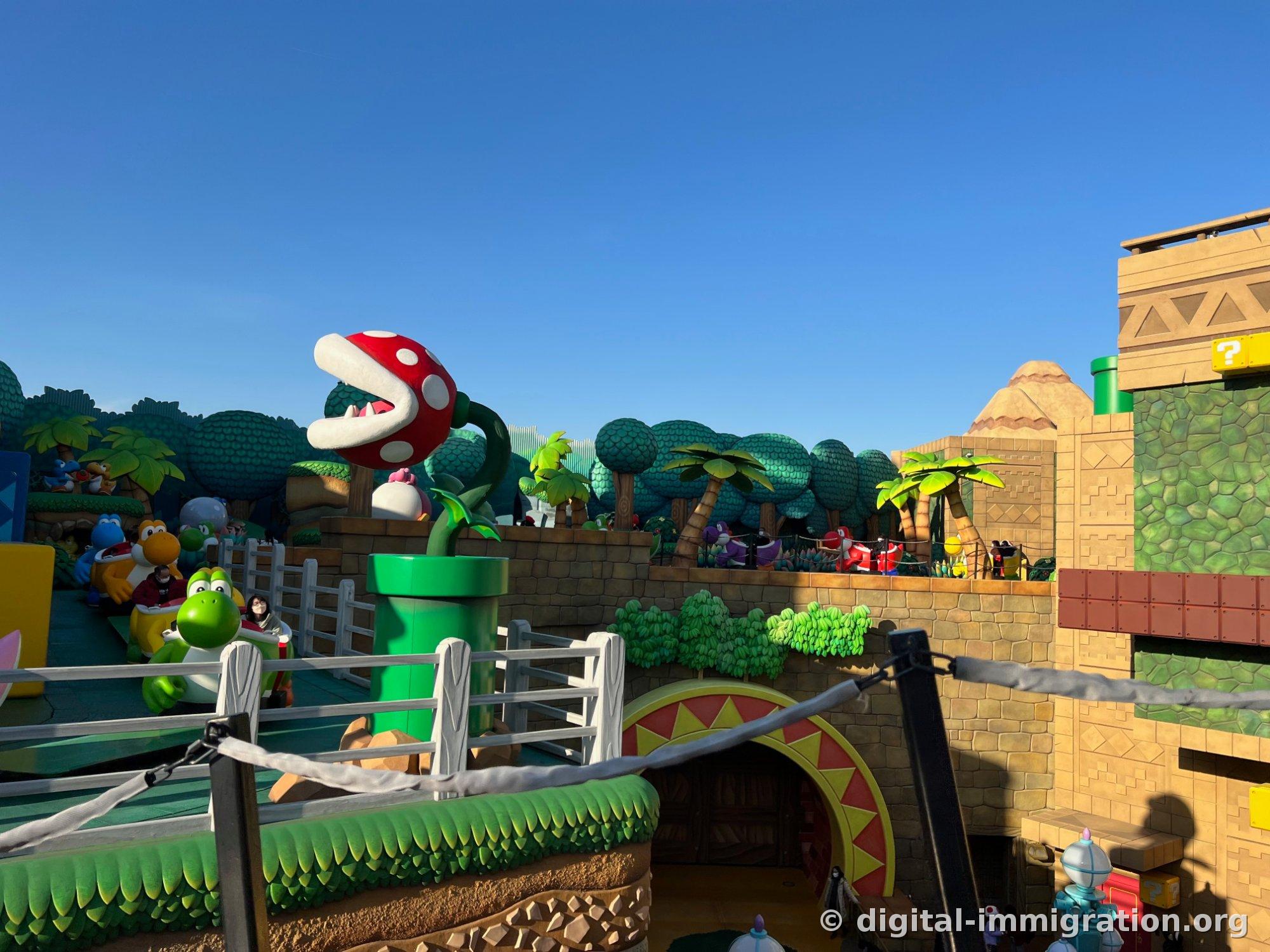 Yoshi Adventure outdoor section. People sit on green Yoshi ride vehicles, with a large Piranha Plant, desert-style stone wall, and palm tree sculptures lined up ahead