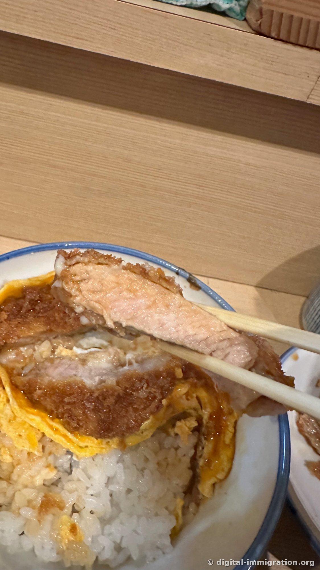 Une baguette soulevant un morceau de katsudon. L'extérieur est de la panure marron, l'intérieur de la viande avec une teinte rosée légère, et en dessous se trouvent l'œuf et le riz blanc