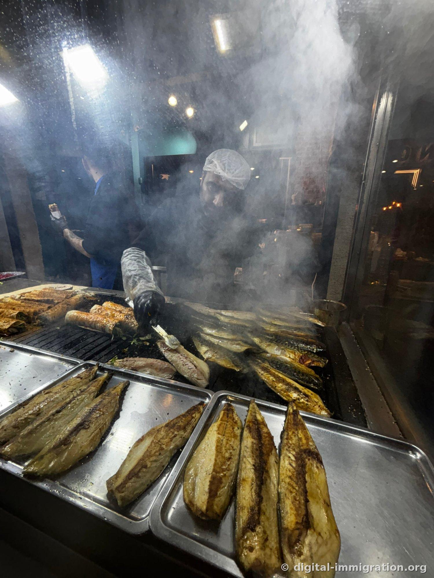 Chef grilling mackerel fillets on an iron plate through a glass window with smoke rising, grilled mackerel stacked in a stainless steel tray in front